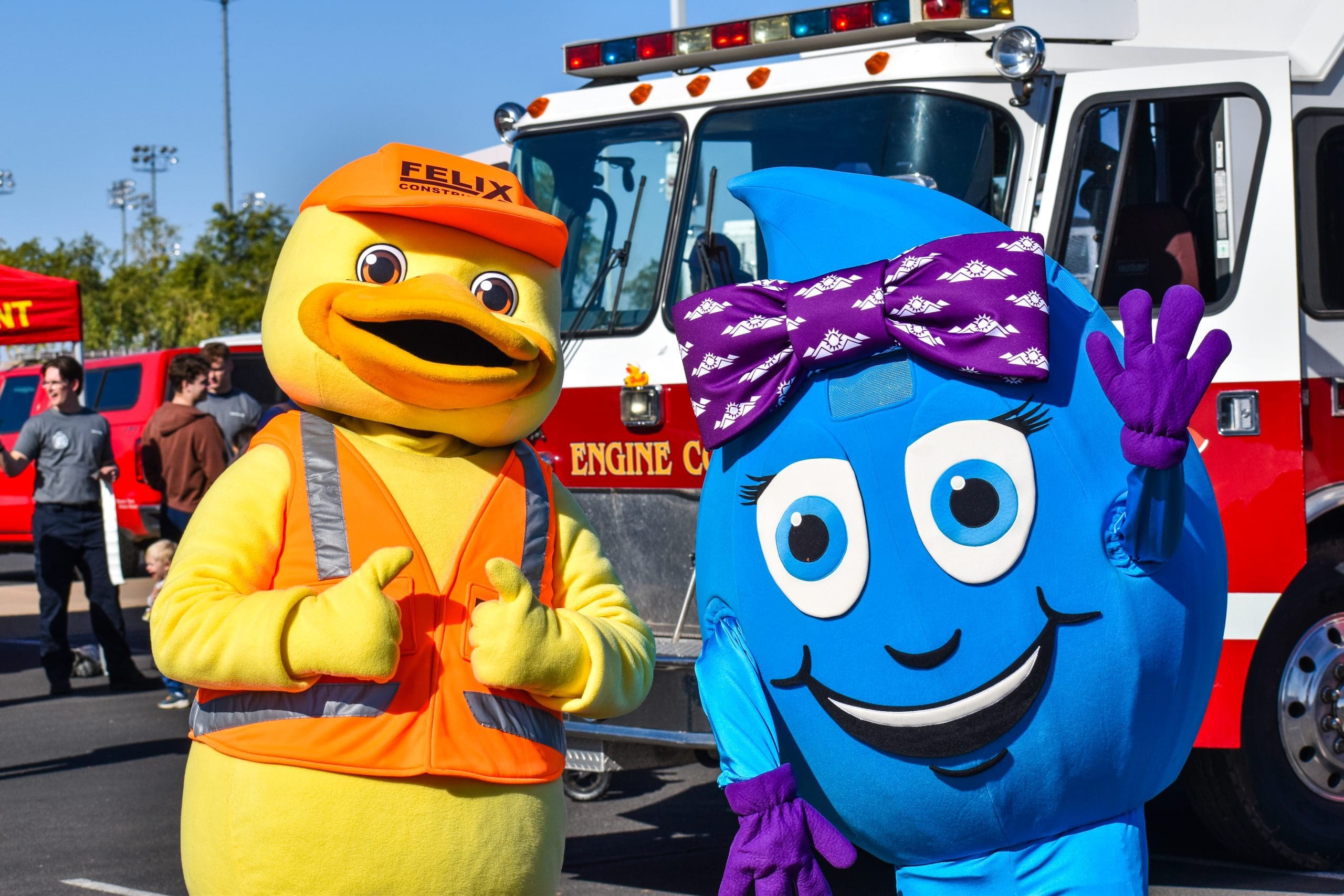A person in a duck costume and another in a blue water droplet costume pose in front of a fire truck at an outdoor event.