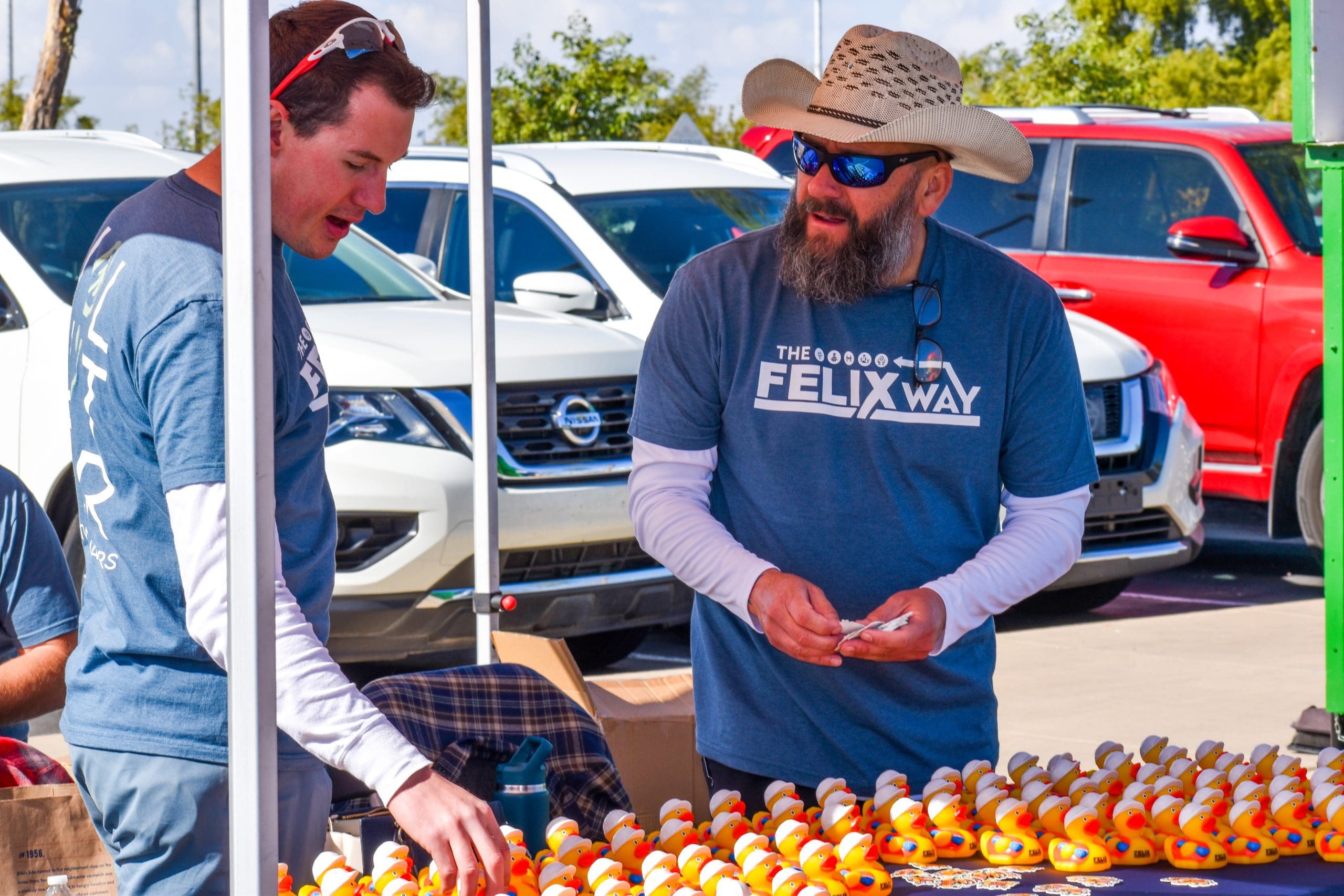 Two men in blue "The Felix Way" shirts stand at an outdoor table covered with rubber ducks, with cars parked in the background.