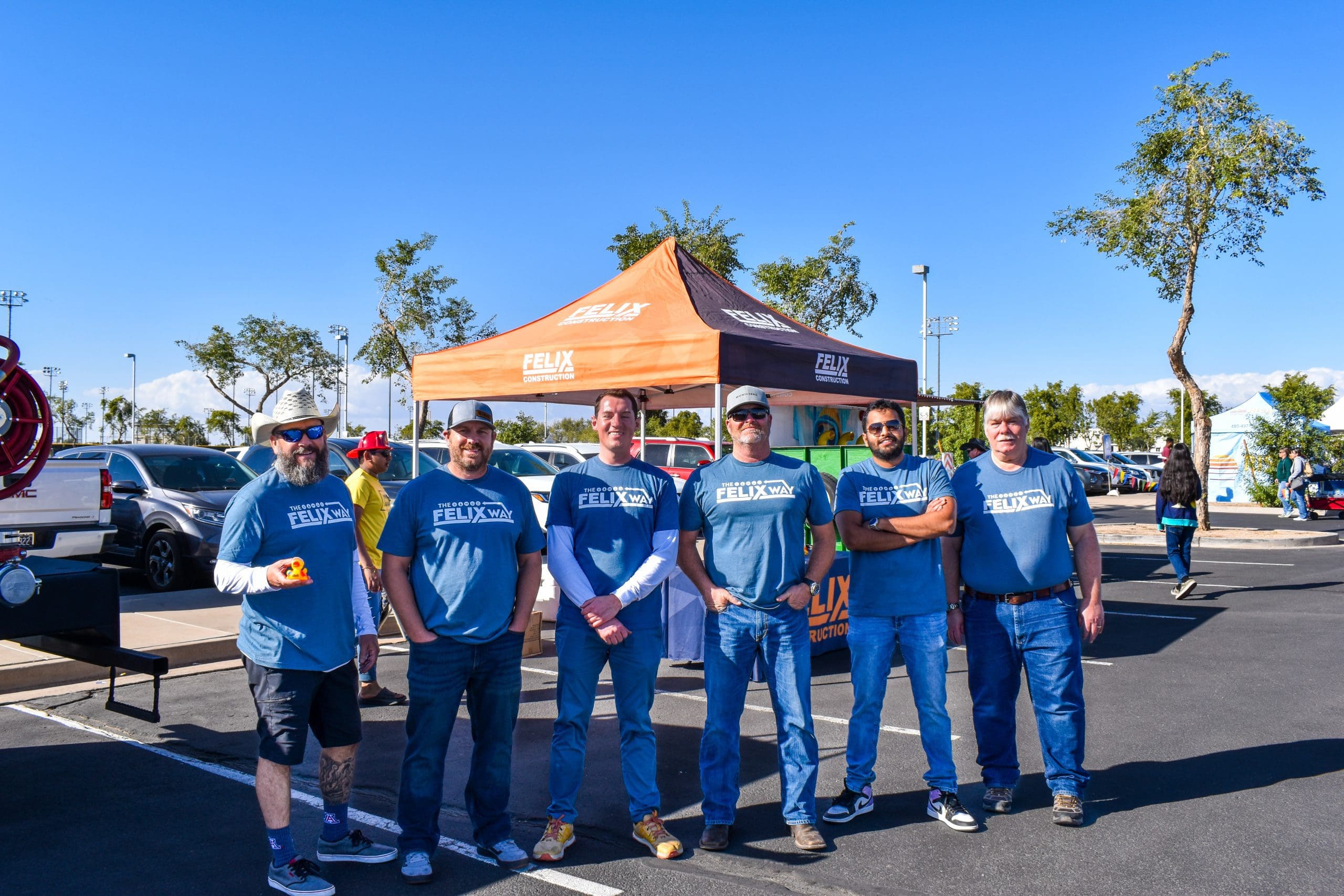 Six men in blue t-shirts stand in a parking lot in front of a branded canopy tent on a sunny day, with vehicles and trees in the background.