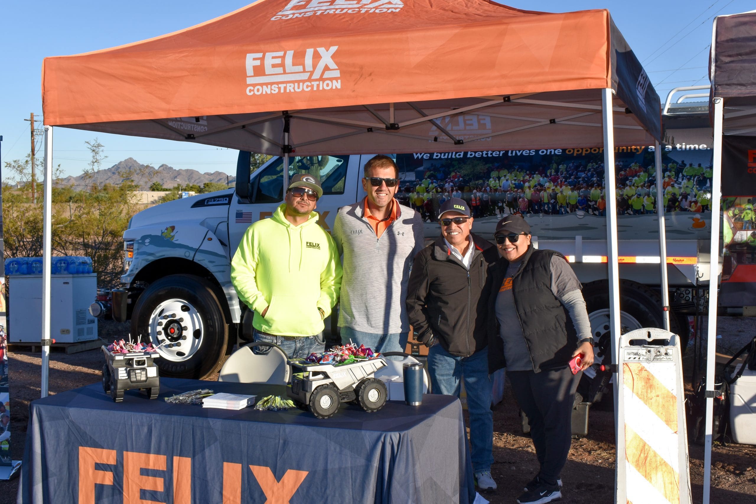 Four people stand under a Felix Construction tent at an outdoor event, with promotional items displayed on a table and a company truck in the background.