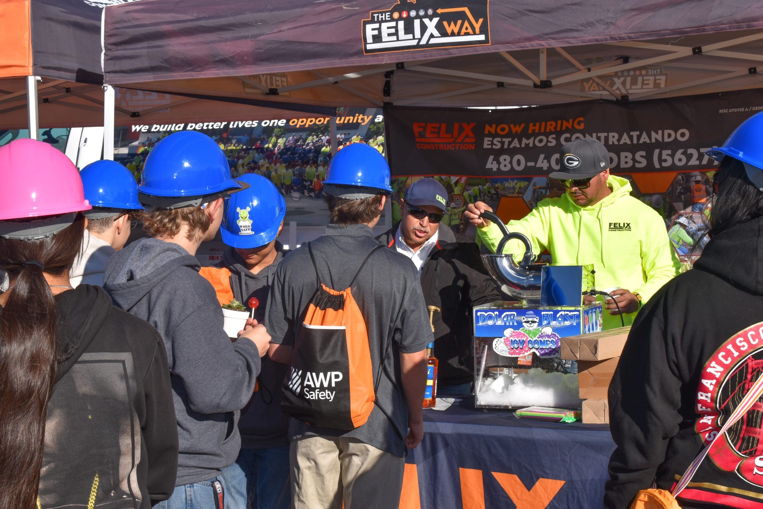 A group of people in hard hats gather at an outdoor booth under a tent, where two men stand behind a table displaying equipment and a "Now Hiring" sign.