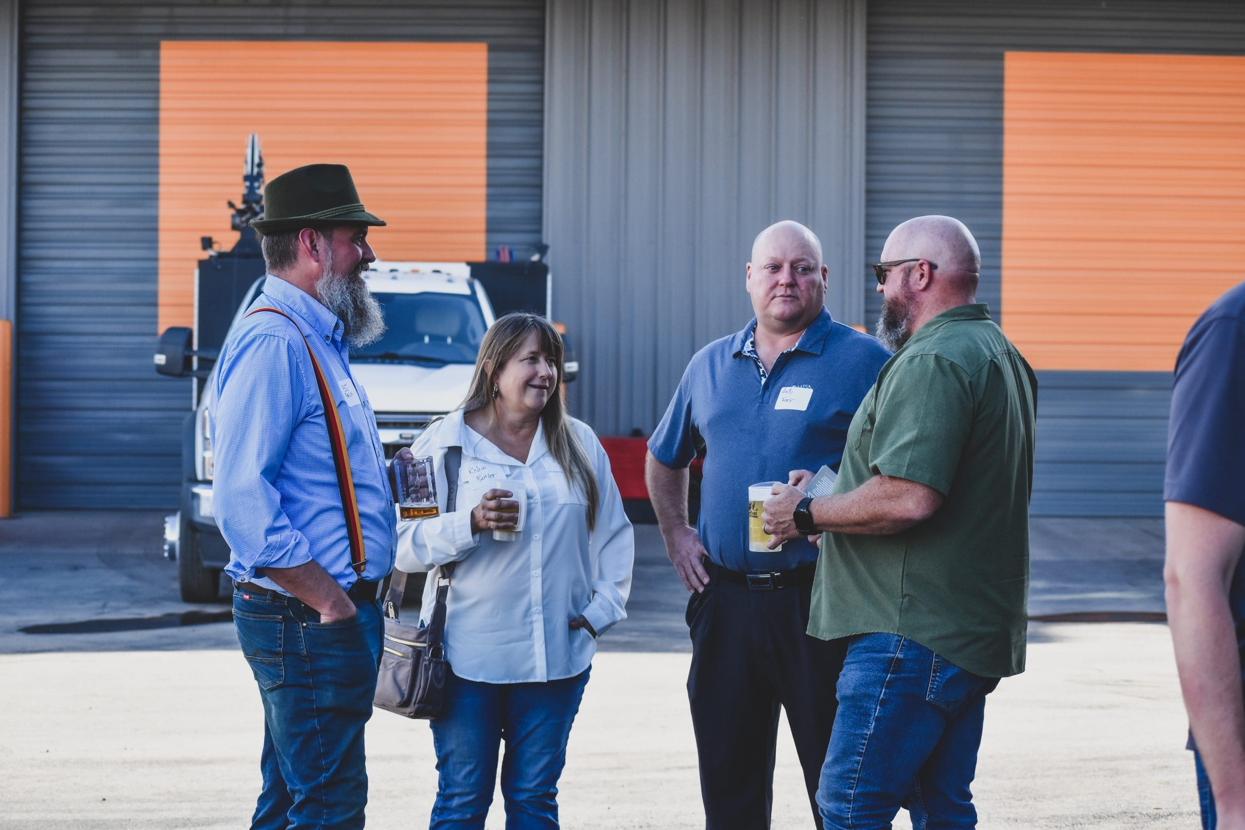 Four adults stand talking and holding drinks in an outdoor industrial area, with a truck and orange-and-gray building visible in the background.