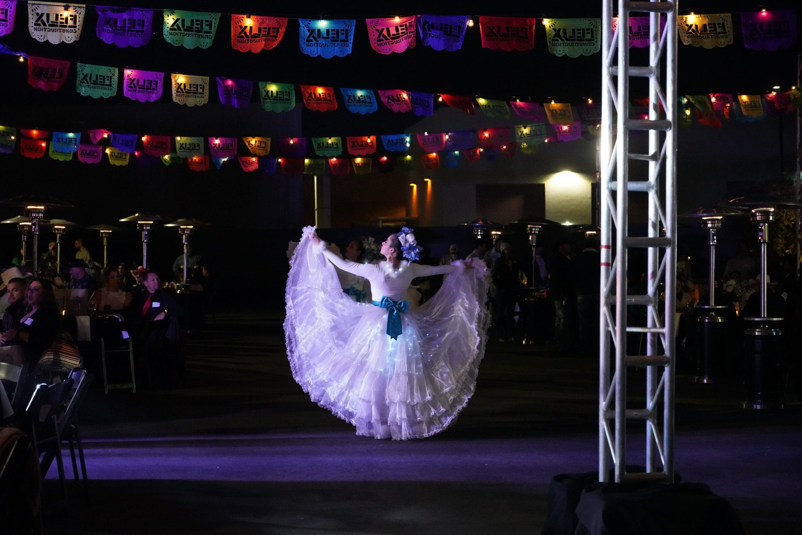 A woman in a white traditional dress performs a folkloric dance on a stage, with colorful papel picado banners hanging above and an audience seated on both sides.