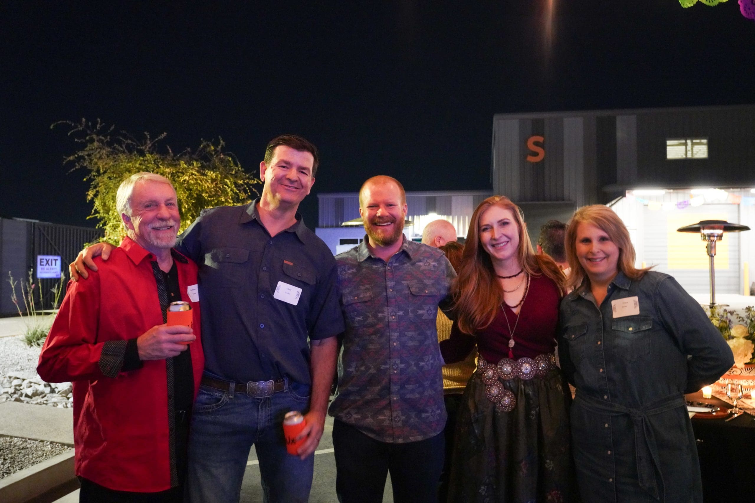 Five adults stand smiling together at an outdoor nighttime event, with name tags visible and buildings in the background.
