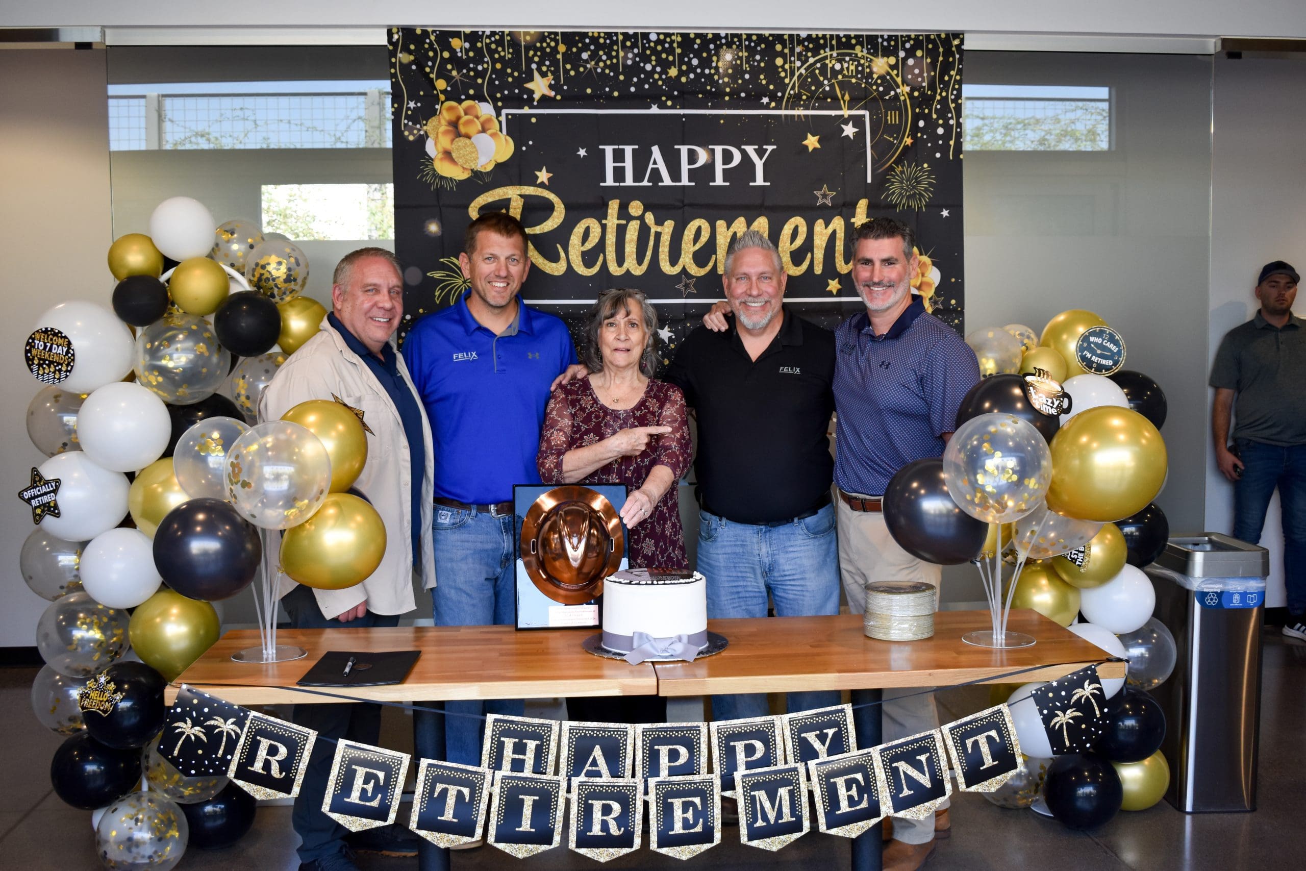 Five people stand in front of a "Happy Retirement" banner and balloons, with a decorated cake and gifts on the table, celebrating a retirement.