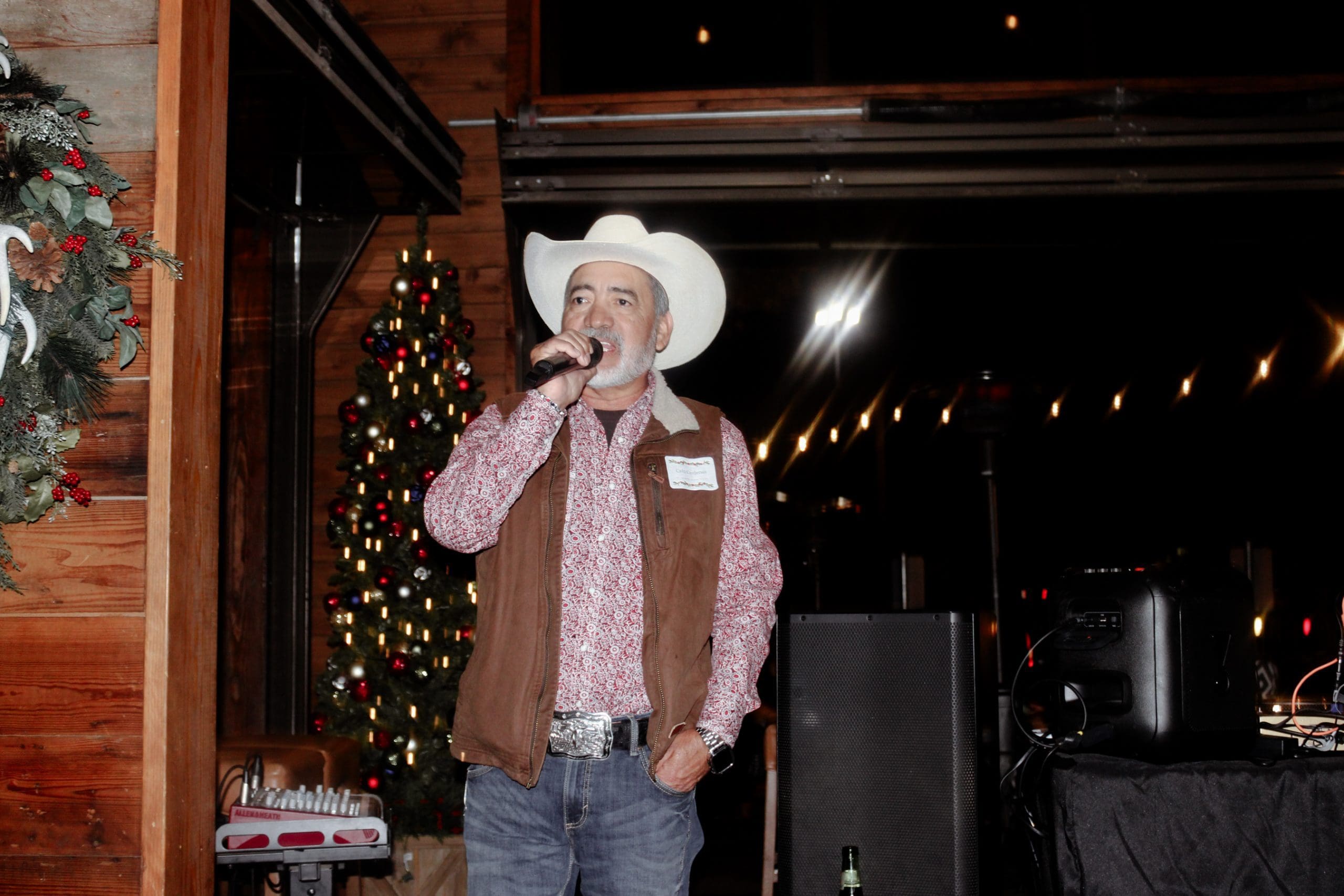 A man in a cowboy hat and brown vest sings into a microphone indoors, with a decorated Christmas tree and audio equipment in the background.