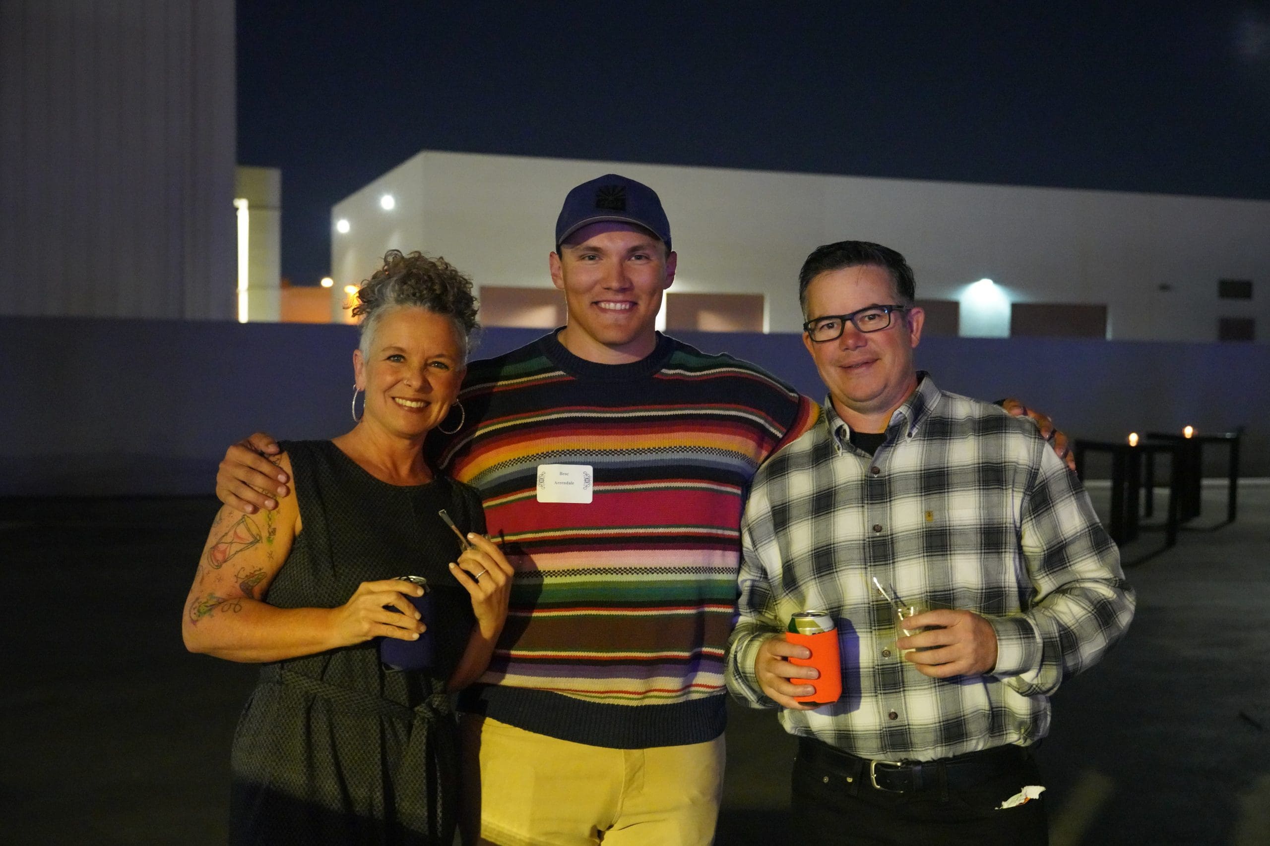 Three adults stand outdoors at night, smiling at the camera. Two hold drinks, and one wears a name badge and a colorful striped sweater. The background shows industrial buildings and minimal lighting.