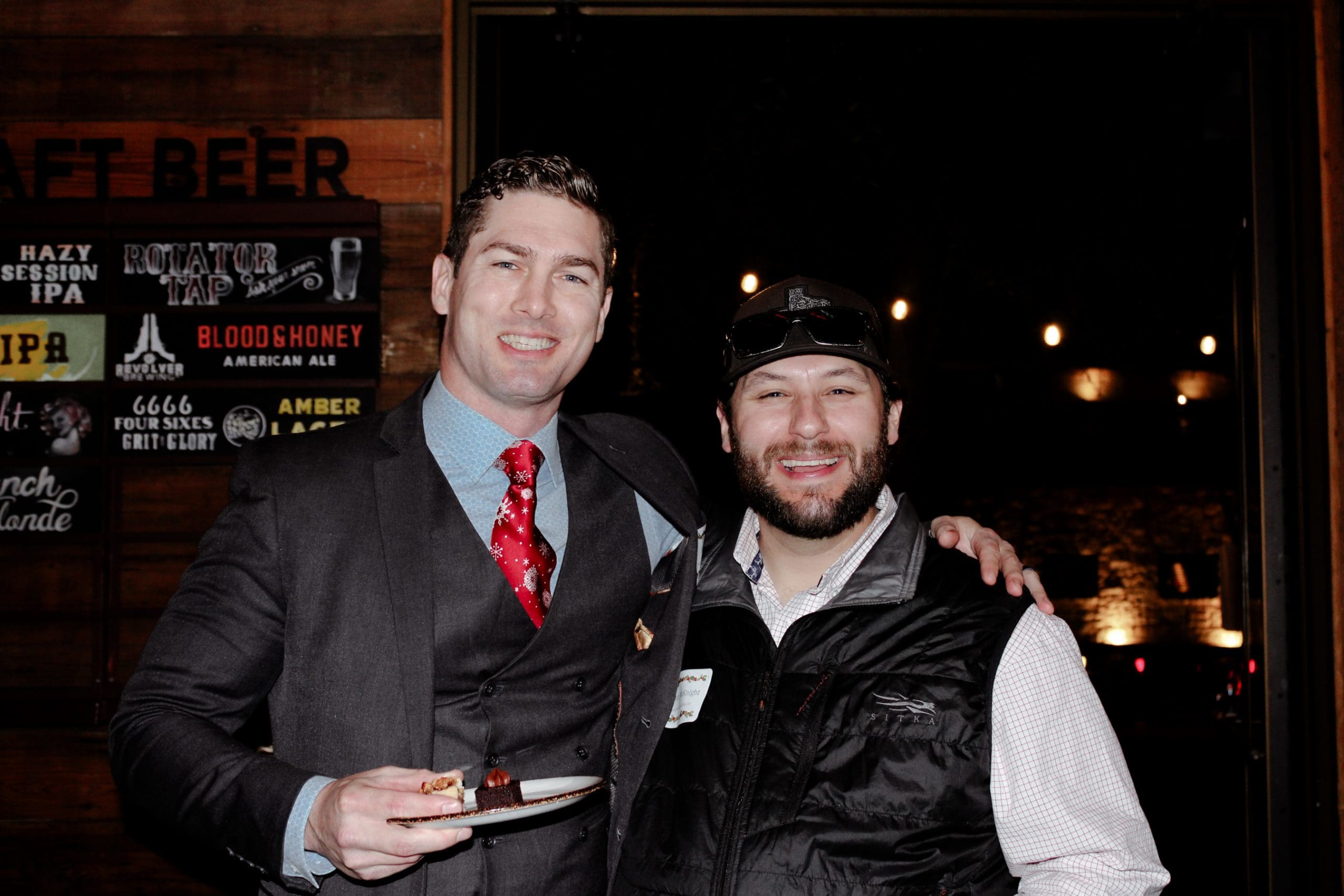 Two men pose and smile for a photo indoors; one is wearing a suit and holding a plate, the other wears casual clothes and a cap. Signs for various beers are visible in the background.