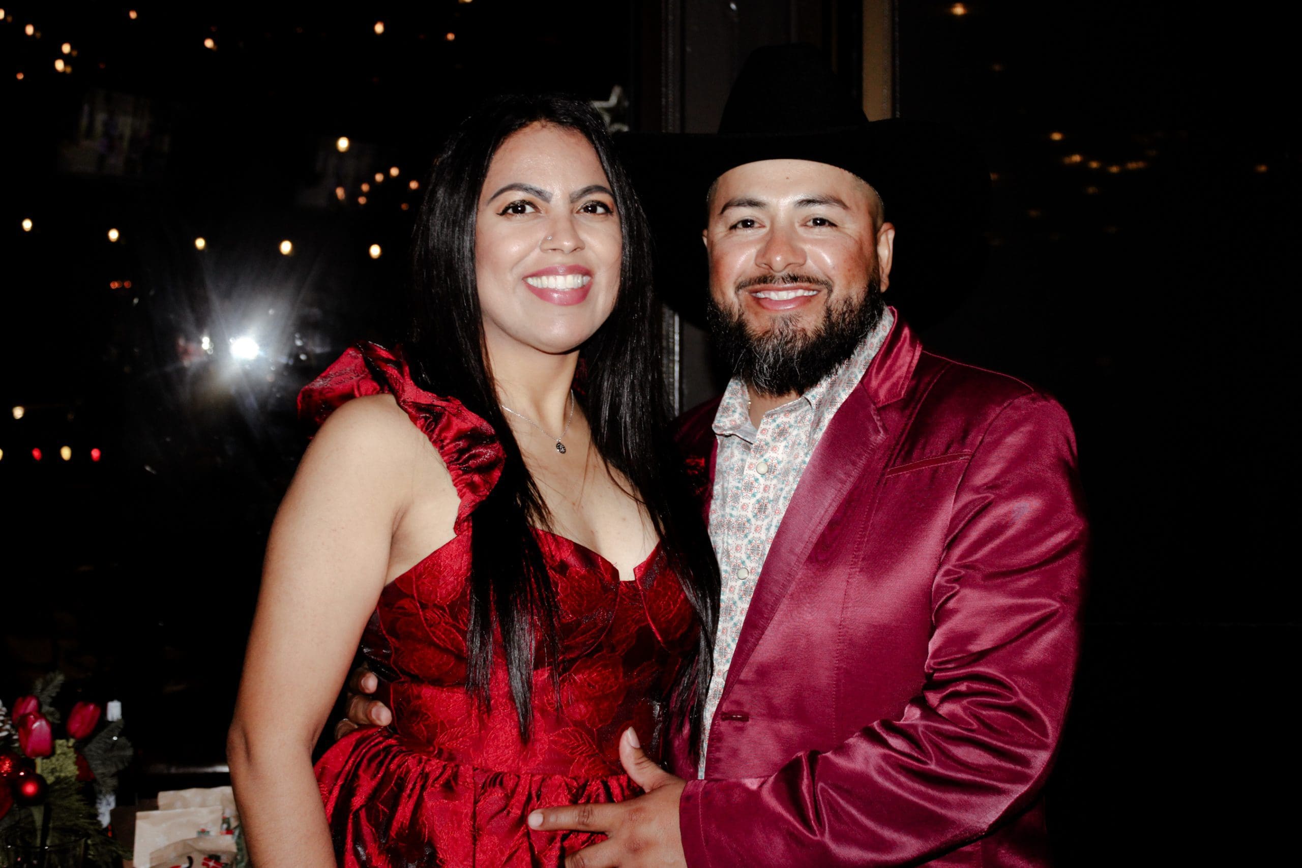 A woman in a red dress and a man in a maroon suit and cowboy hat pose together and smile at the camera in a dimly lit setting.
