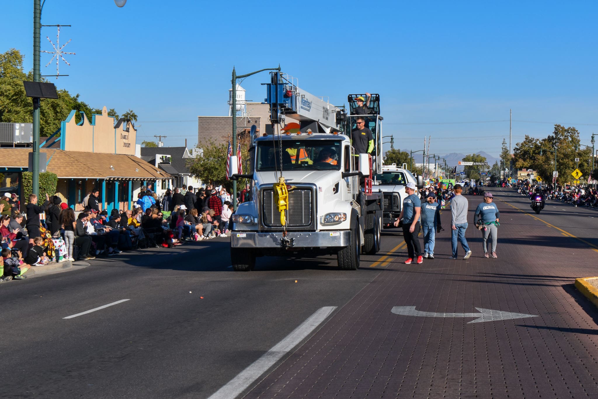 A utility truck drives down a street during a parade, with people sitting and standing along the sidewalks and a few walking near the truck.