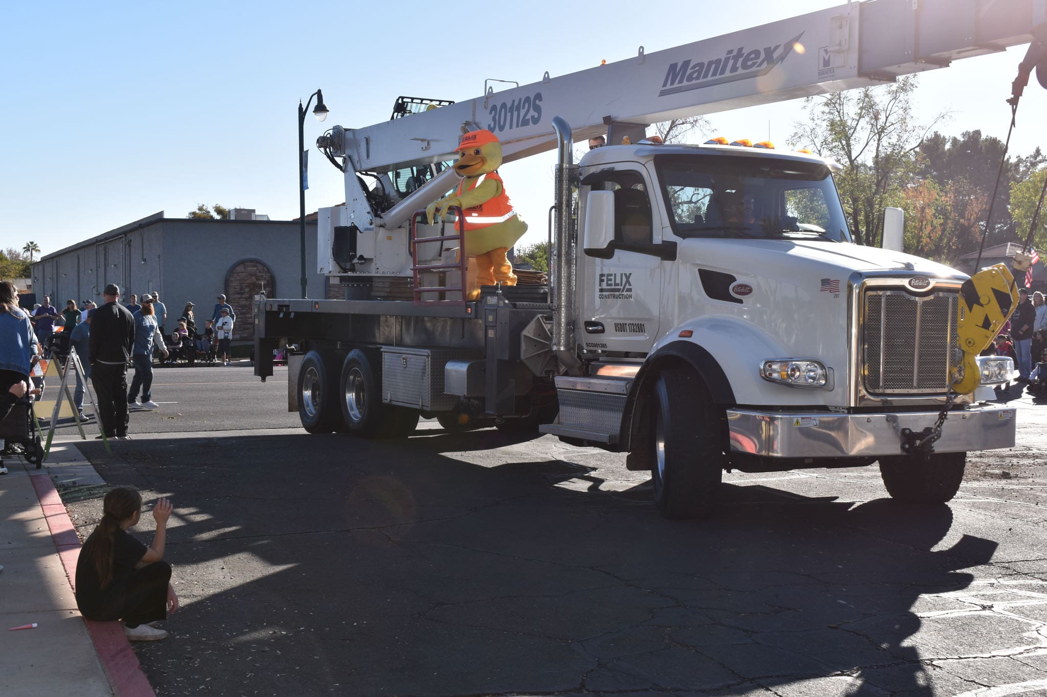 A large white crane truck drives in a parade; a person in a duck costume wearing a safety vest stands on the truck. Spectators watch from the sidewalk.