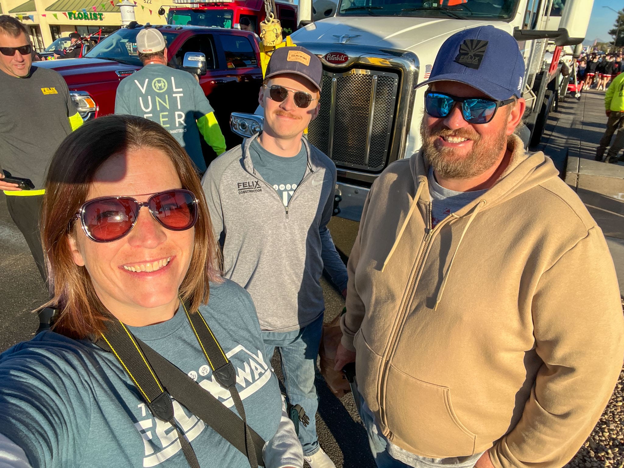 Three adults wearing sunglasses and casual clothes stand outdoors smiling at the camera, with trucks, people, and a volunteer in the background.