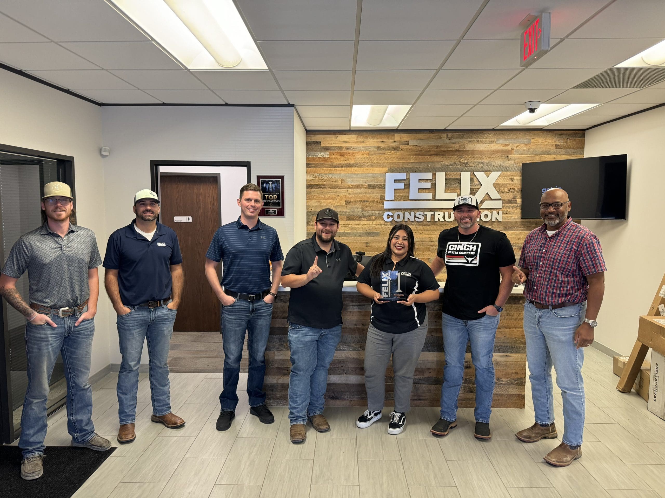 Seven people stand and smile in front of a wooden wall with a "FELIX CONSTRUCTION" sign in an office lobby. One person holds a plaque or award.
