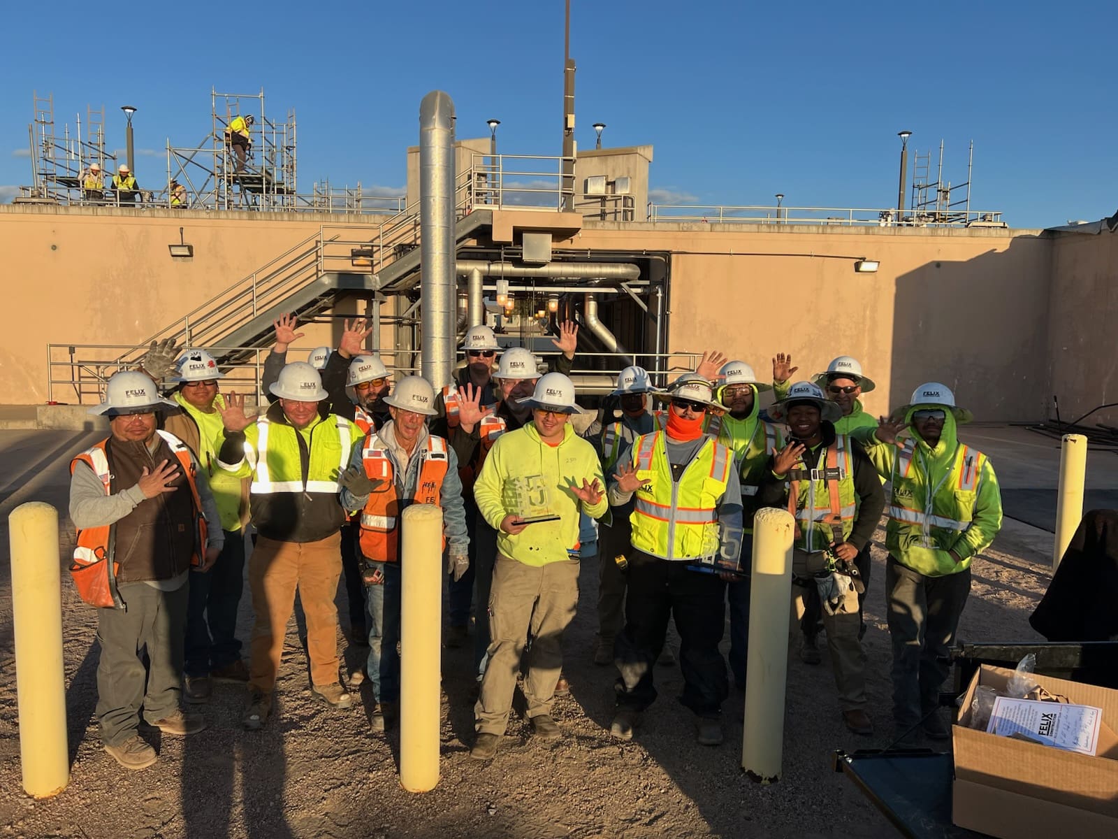 A group of construction workers in safety vests and helmets pose together outside an industrial facility, holding up their hands and smiling at the camera.