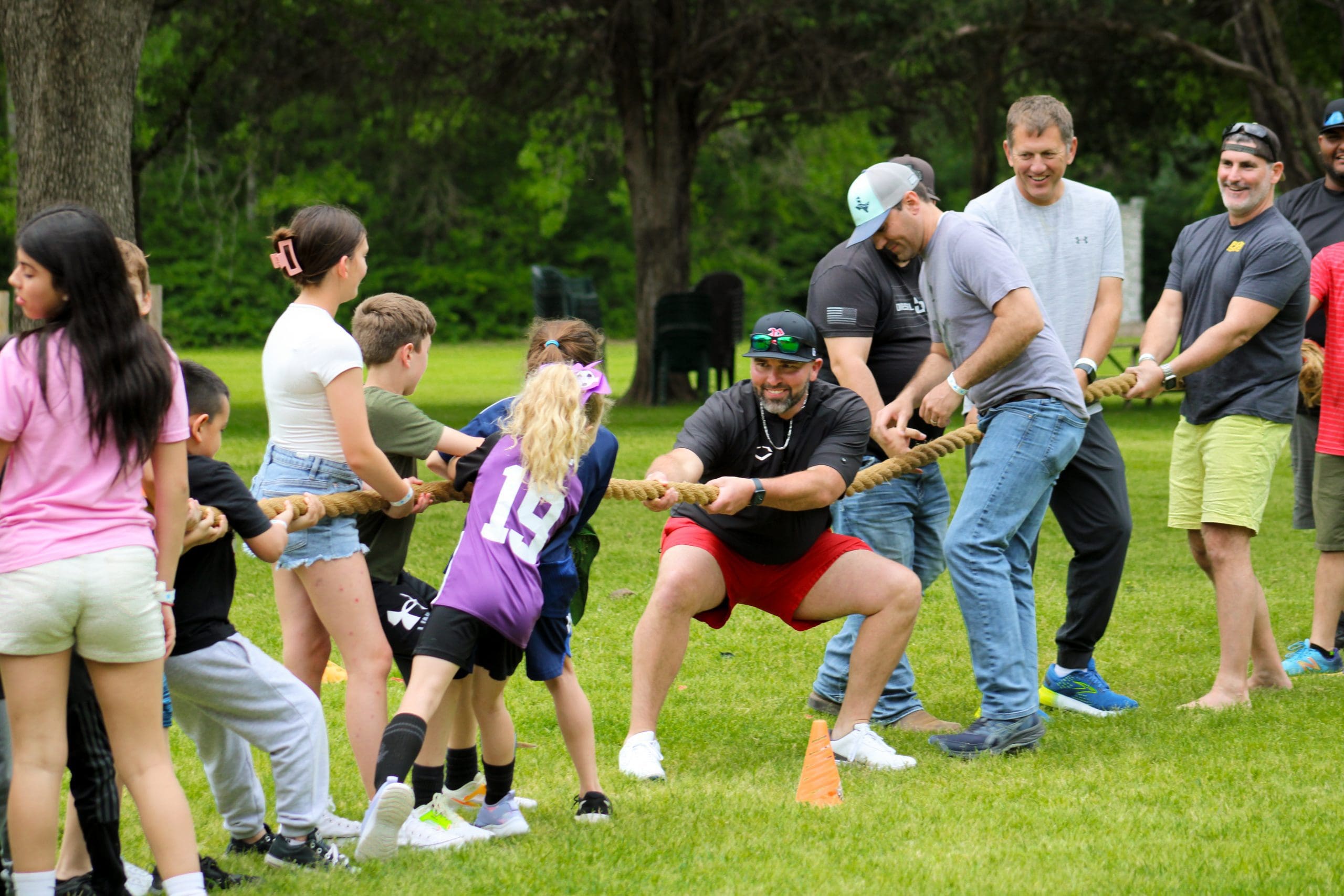A group of adults and children play tug-of-war on a grassy field, pulling on a rope and smiling, with trees in the background.