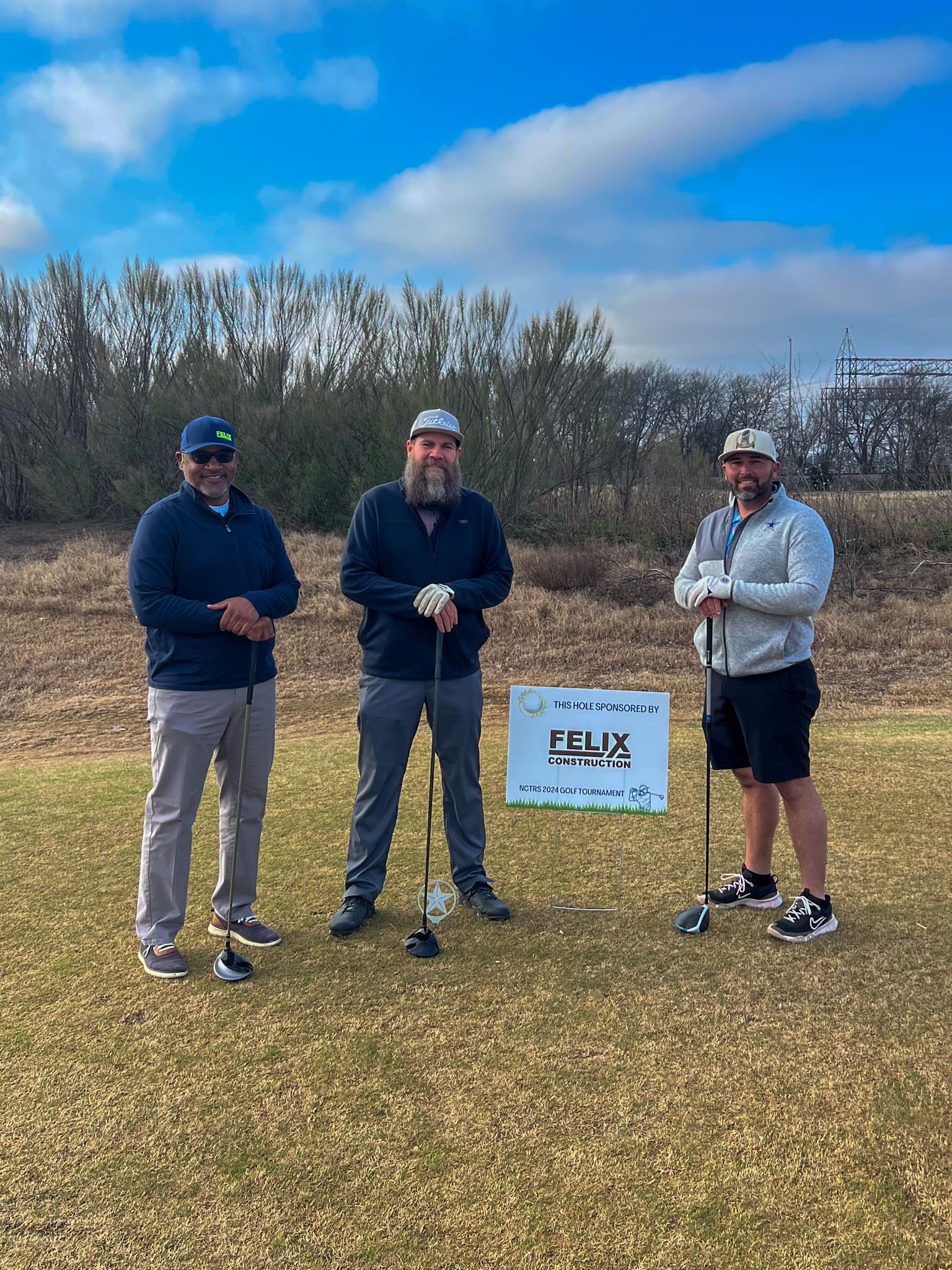 Three men stand on a golf course holding clubs, posing in front of a sign that reads "This hole sponsored by Felix Construction." Dry grass and overcast sky are in the background.