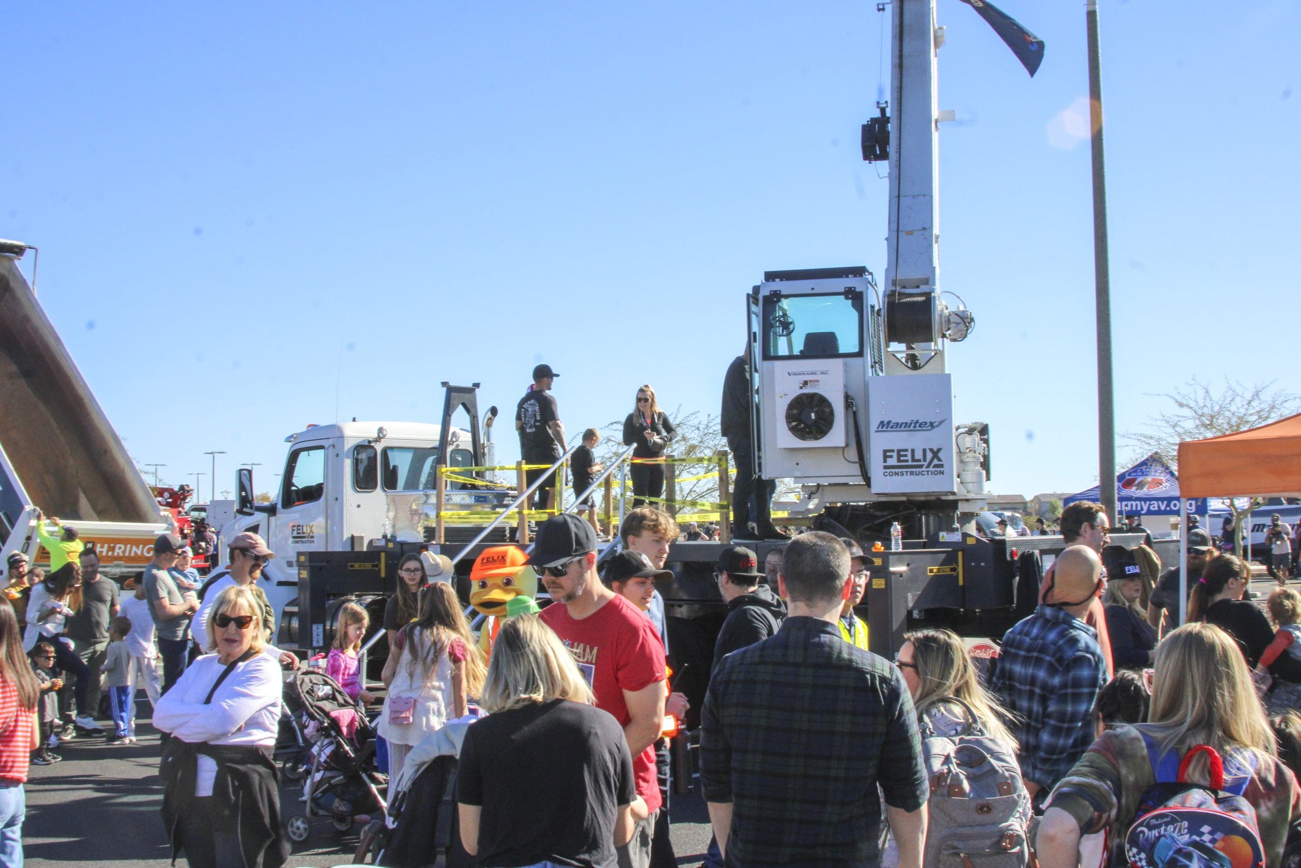 A crowd of people gathers around a large white truck with a crane at an outdoor event on a sunny day. Some individuals are standing on the truck’s platform.