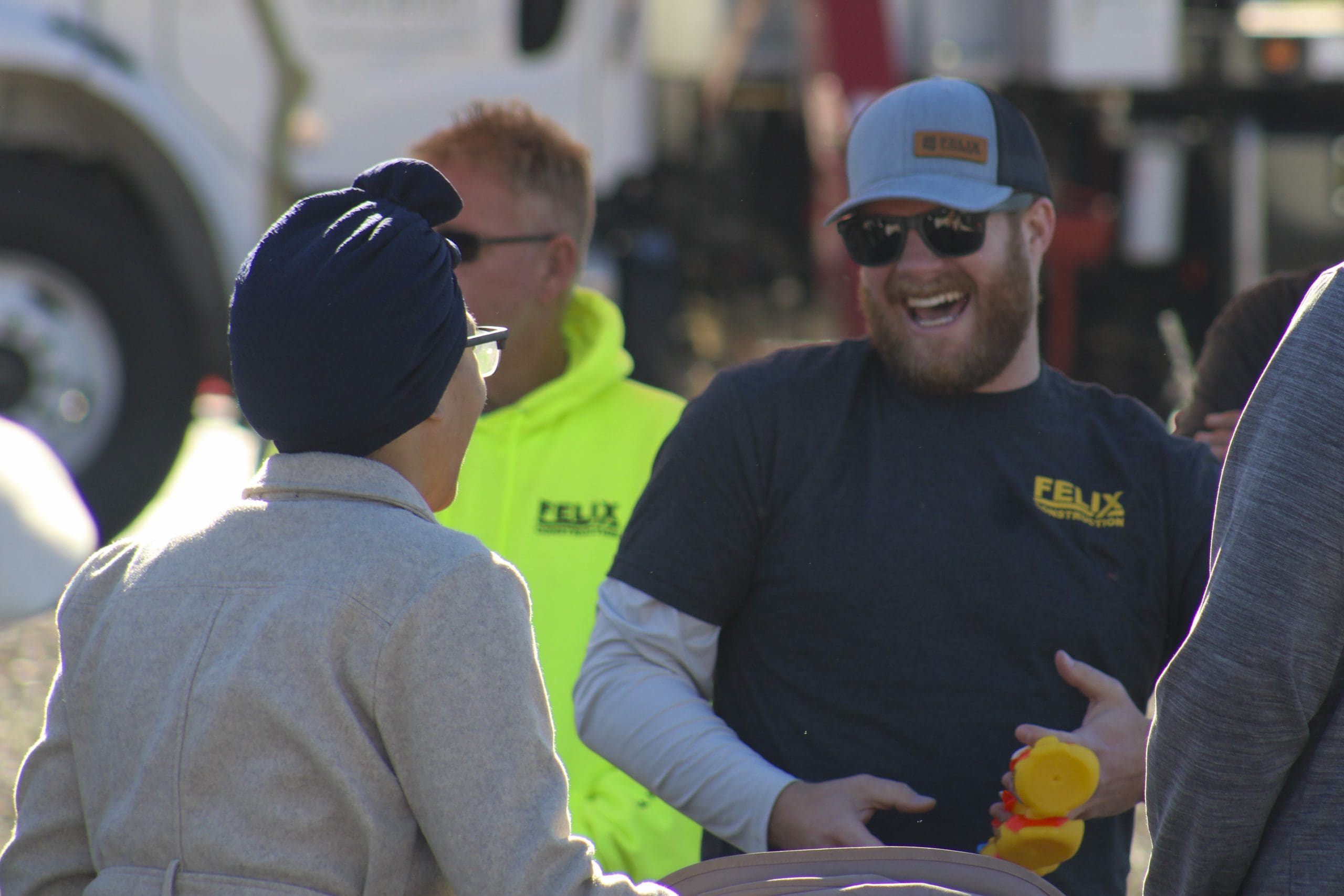 A man wearing sunglasses and a hat laughs while holding a rubber duck, standing with others at an outdoor event.