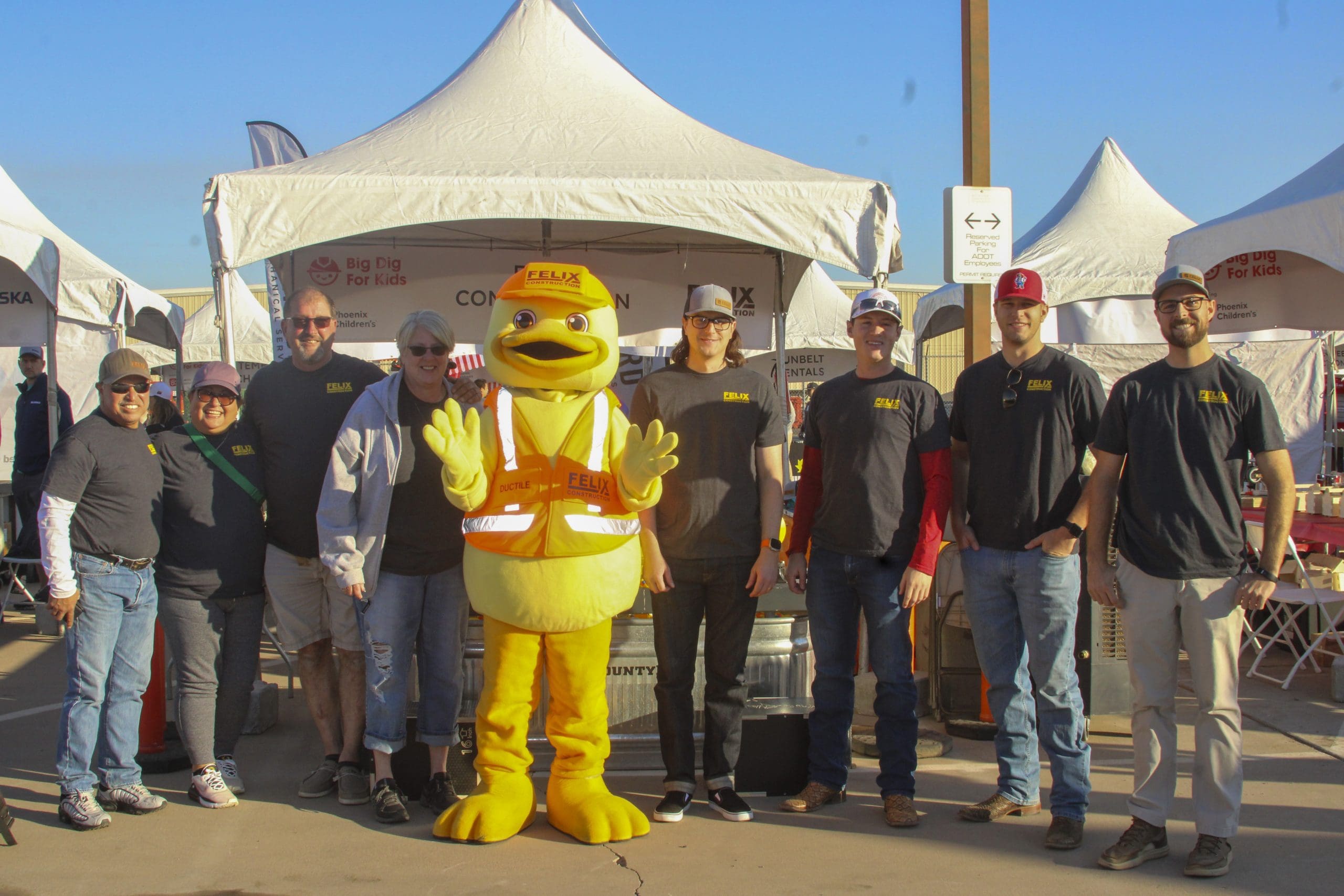 A group of eight people stands on either side of a person in a yellow duck mascot costume in front of white event tents outdoors.