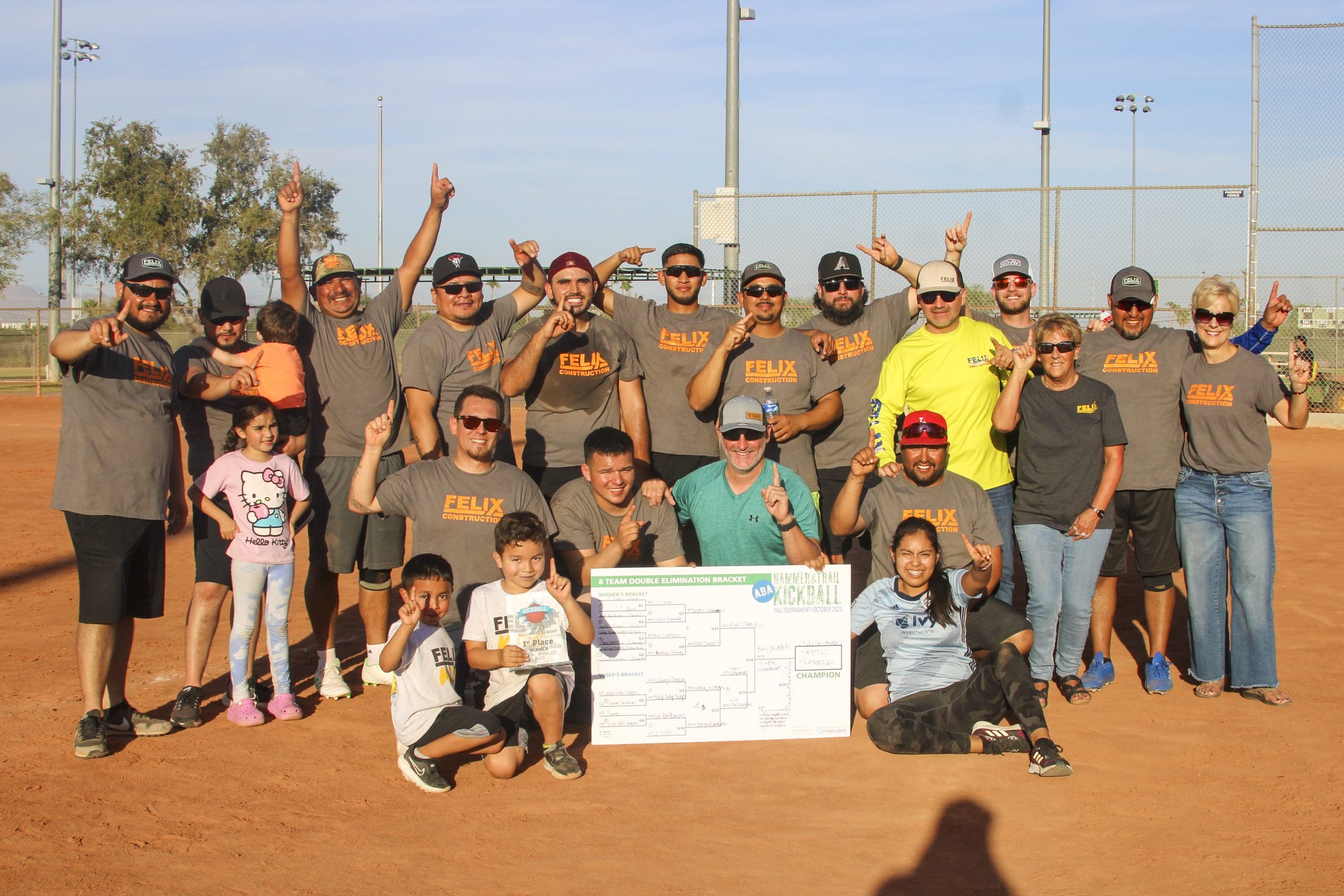 A group of adults and children pose on a baseball field, some holding up one finger, with a large tournament bracket board displayed in front.