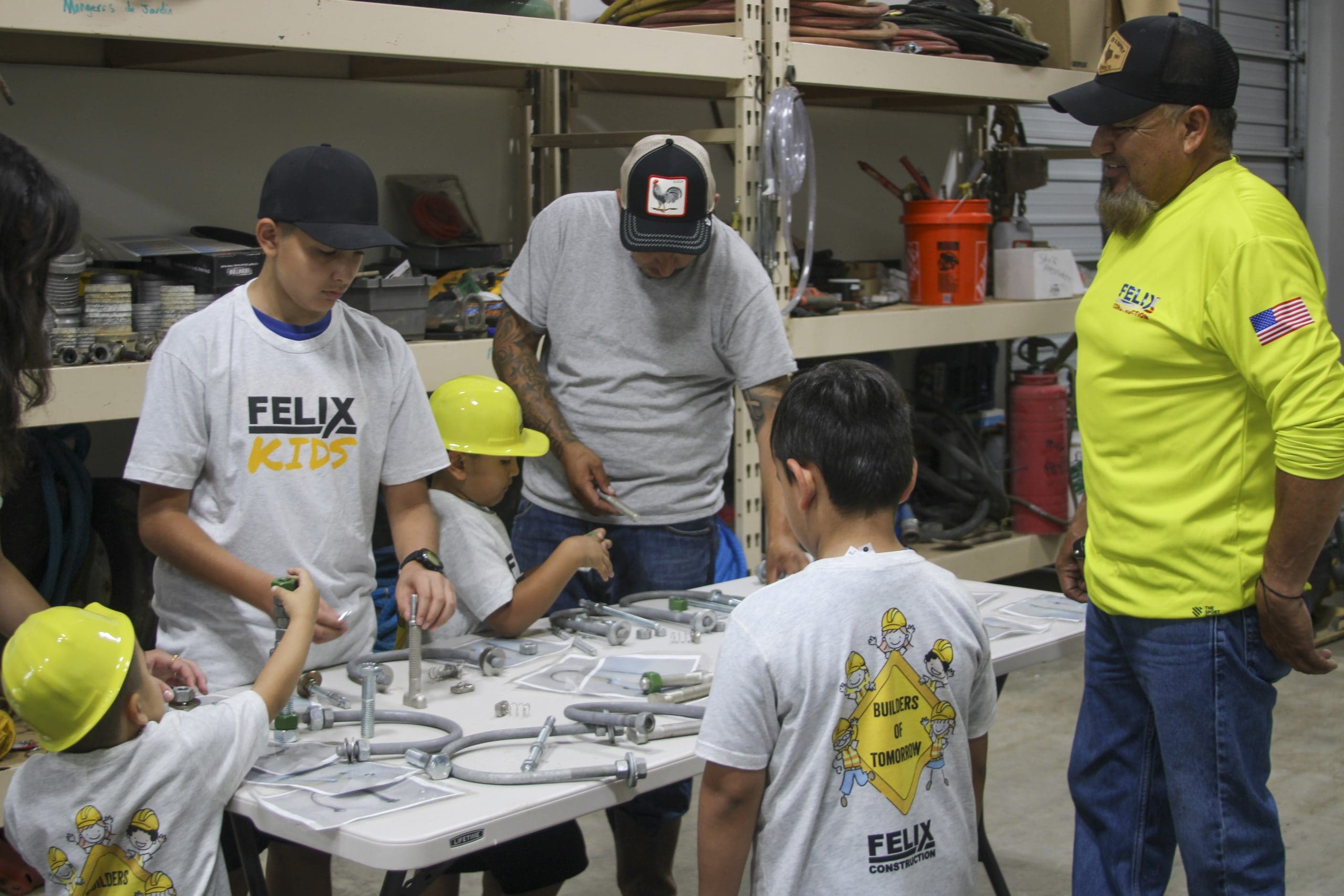Several children and adults work together assembling metal pipe parts on a table in a workshop. Some children wear yellow hard hats and everyone wears Felix branded shirts.