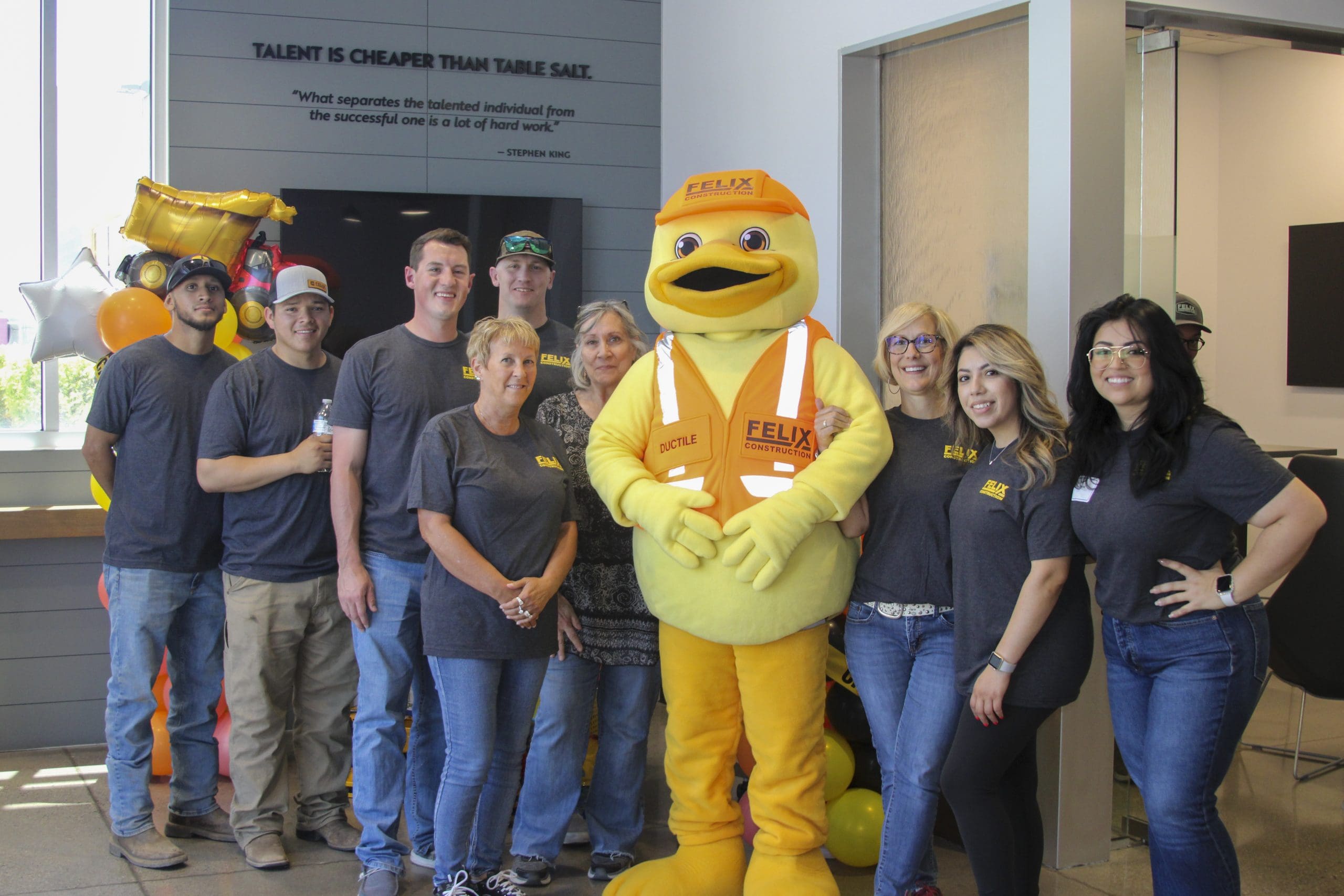 A group of people poses indoors with a person in a yellow duck mascot costume wearing a construction vest and helmet. Balloons and decorations are in the background.