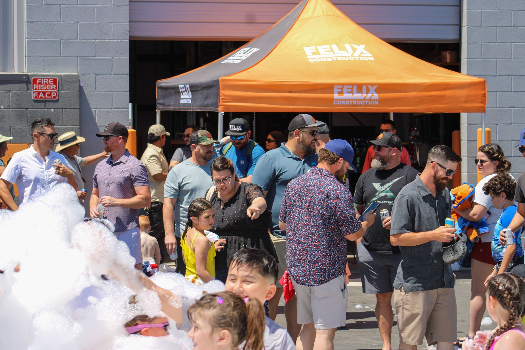 Adults and children gather outdoors near an orange "Felix Construction" tent, with some kids playing in foam in the foreground.