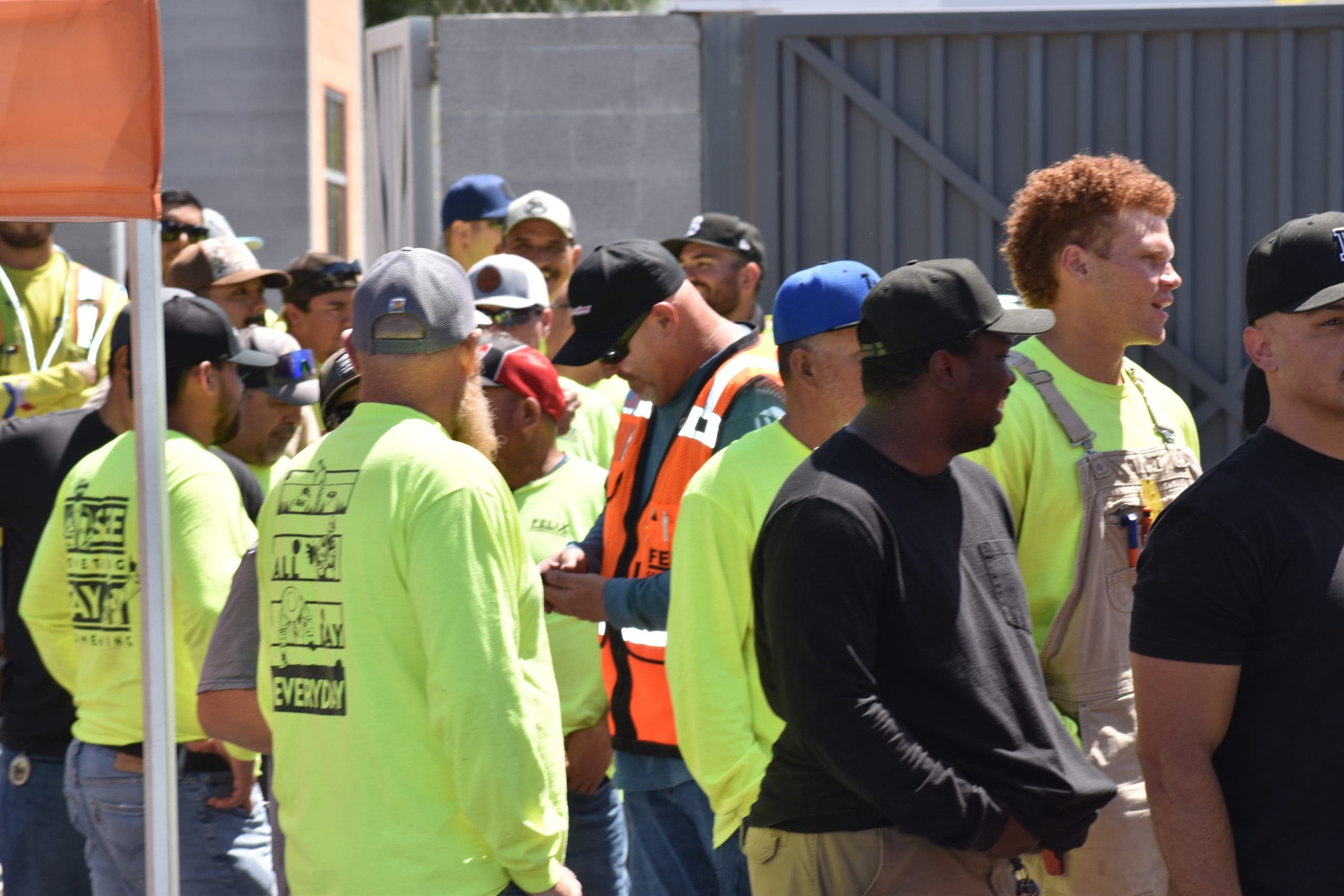 A group of construction workers wearing safety vests and hard hats stand together outdoors near a metal fence and building.
