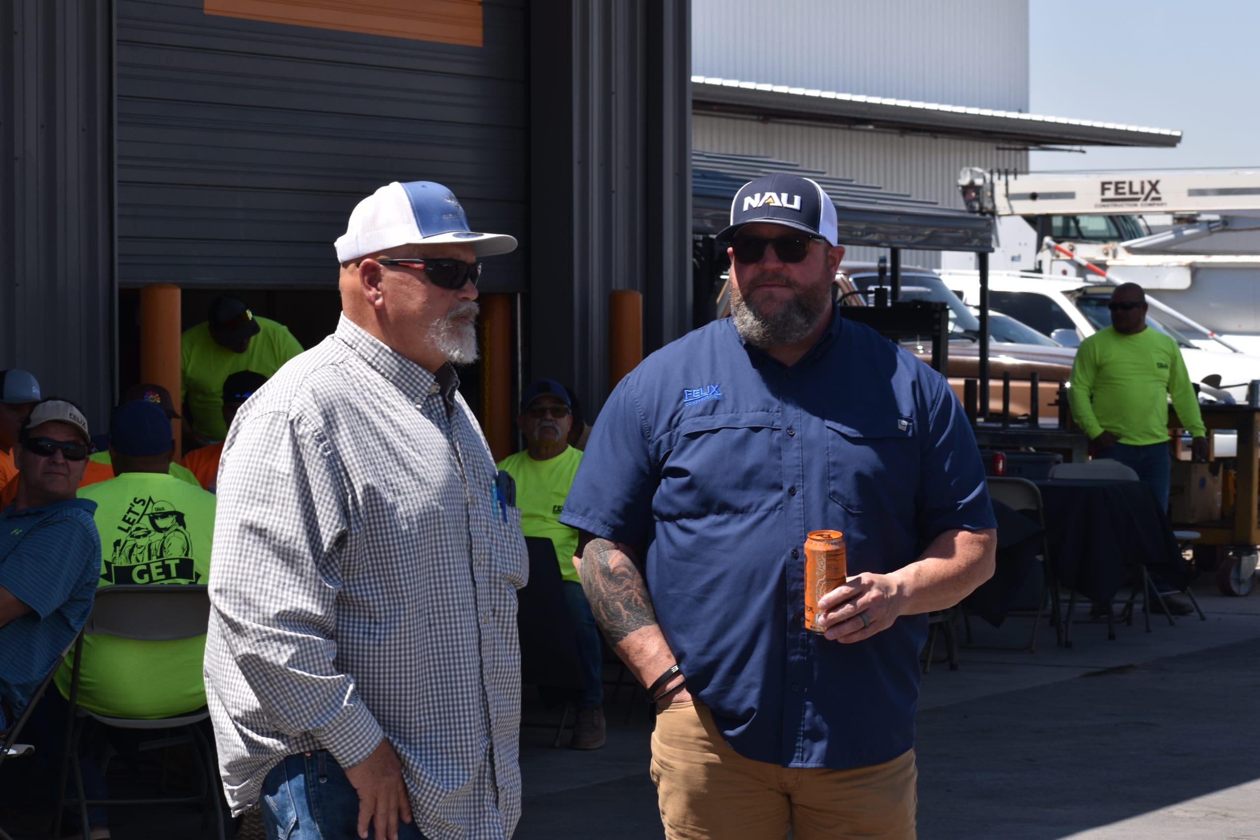 Two men wearing sunglasses and hats stand outside a building, one holding a drink can. Several people in safety vests sit in the background near tables.