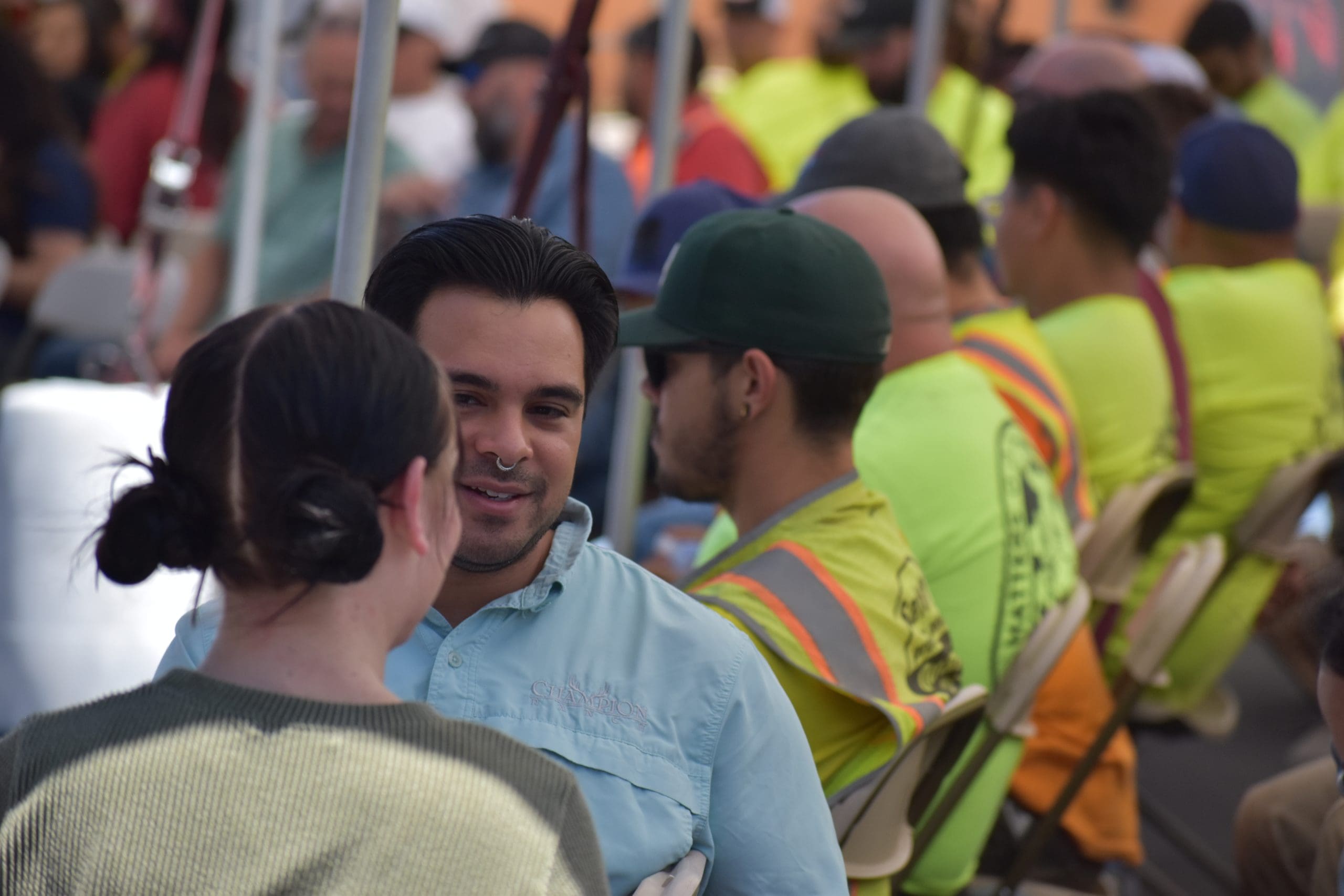 A man and woman converse in the foreground while people in safety vests sit in rows of chairs at an outdoor event.