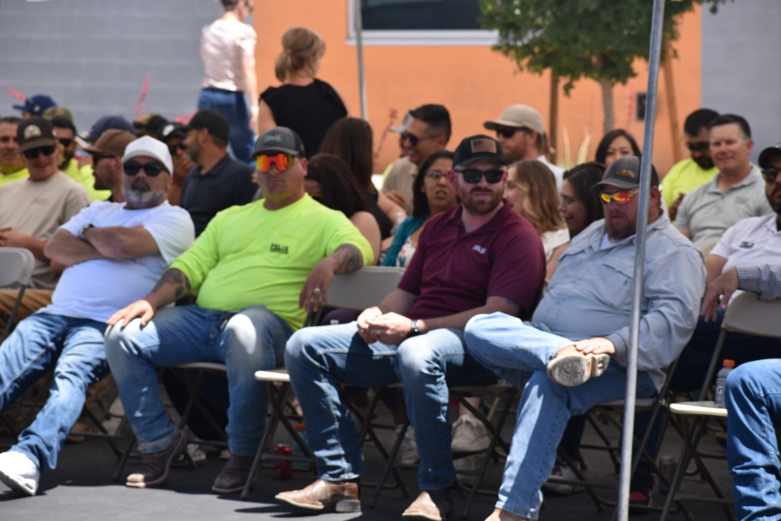 A group of people, mostly men, sit on folding chairs outdoors at an event. Some wear sunglasses and brightly colored shirts. The background includes more attendees and a building.