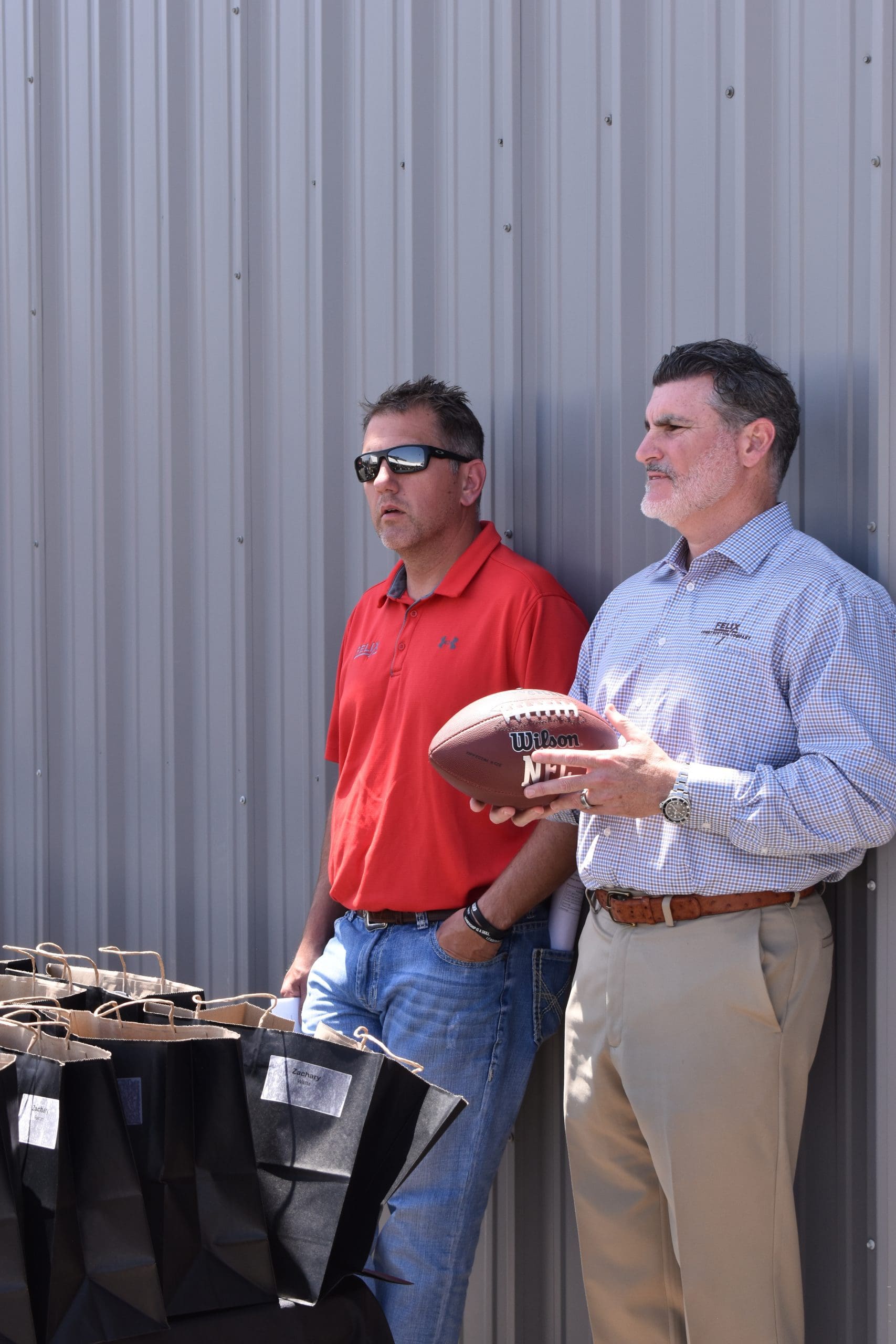 Two men stand beside a metal wall, one holding a football. There are several black gift bags on the table next to them.