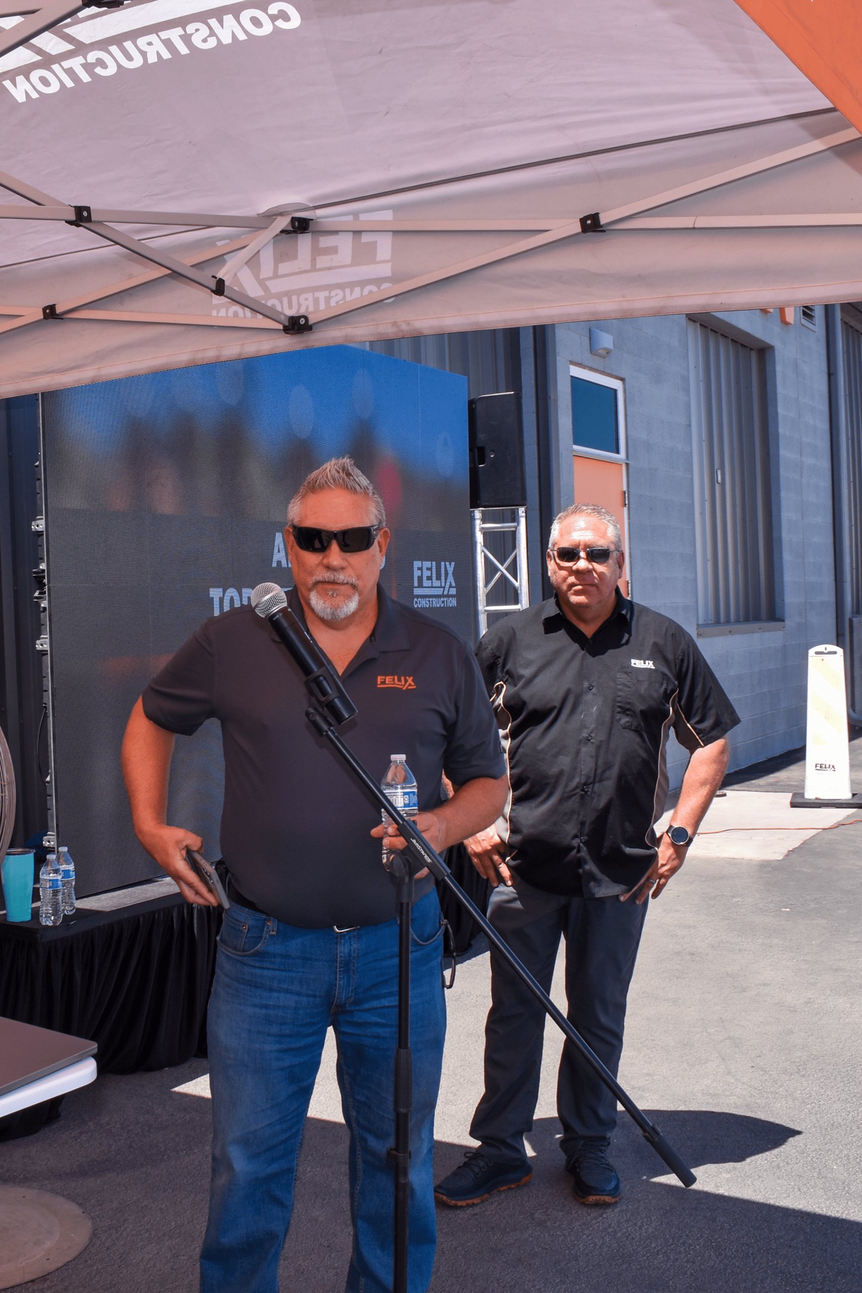 Two men wearing sunglasses stand under a tent at an outdoor event. One man speaks at a microphone, while the other stands behind him near a building.