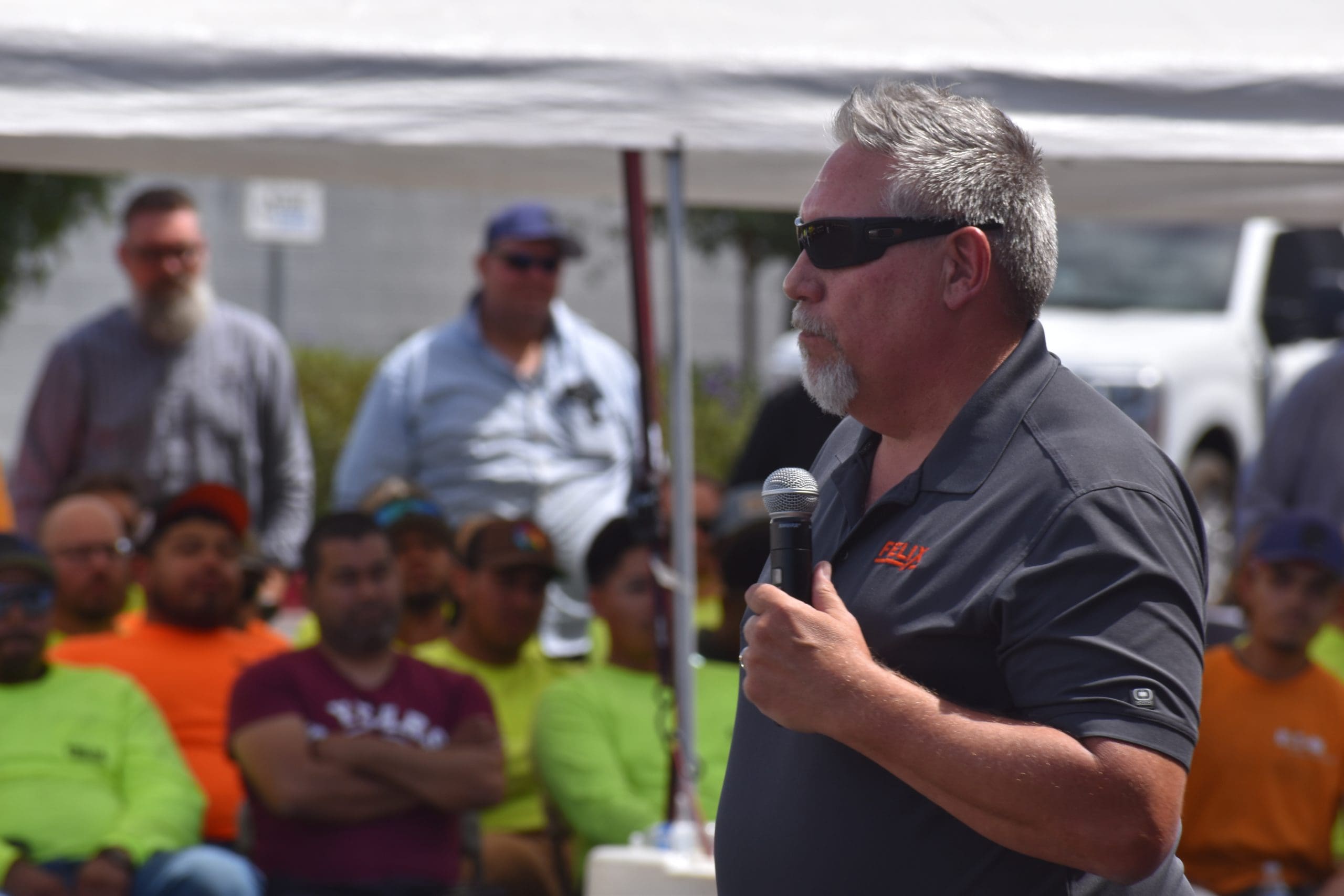 A man in sunglasses speaks into a microphone outdoors to a seated group of people wearing bright shirts, with others standing in the background under a canopy.