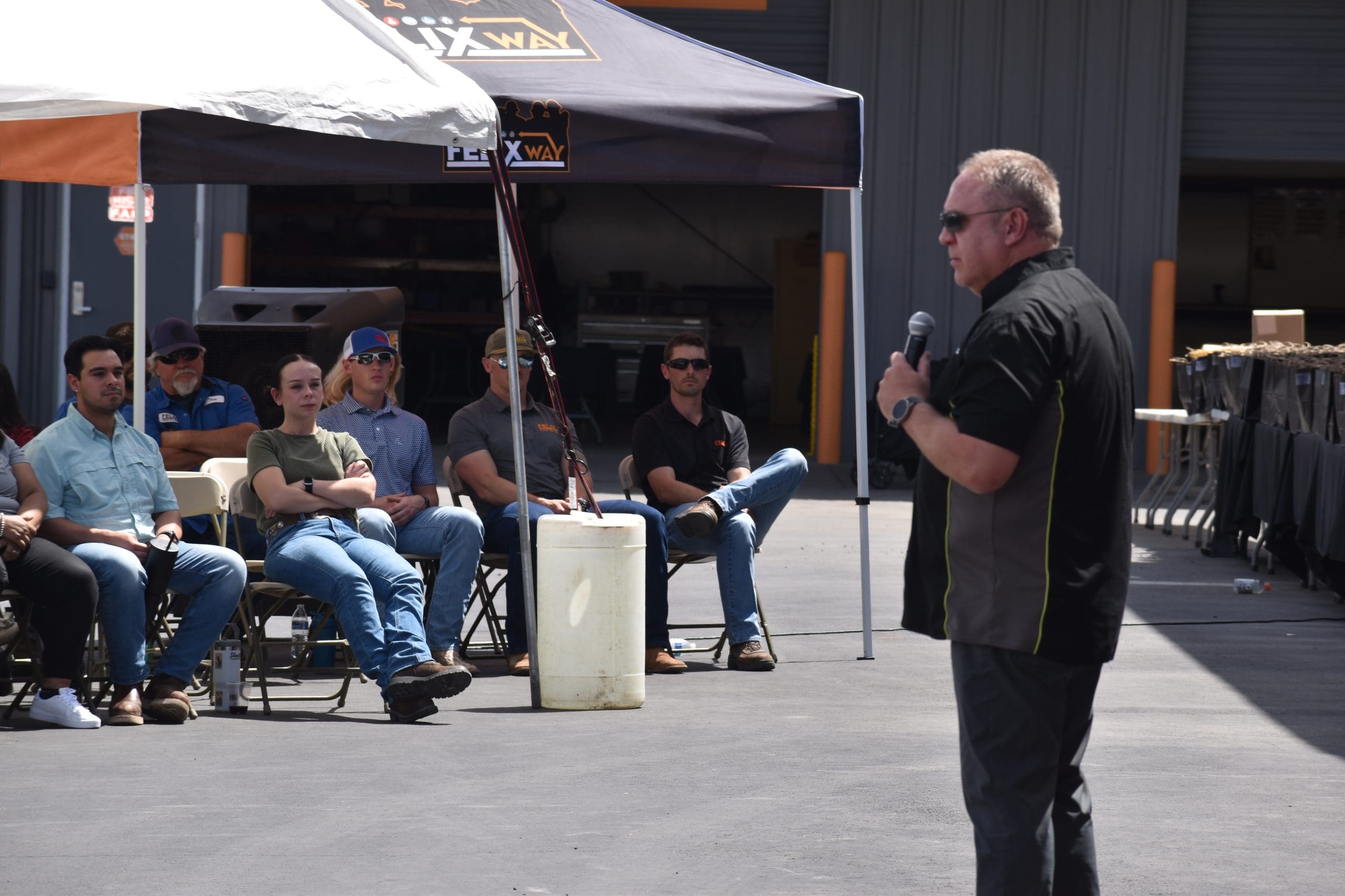 A man speaks into a microphone to a seated audience outdoors under tents in an industrial area.