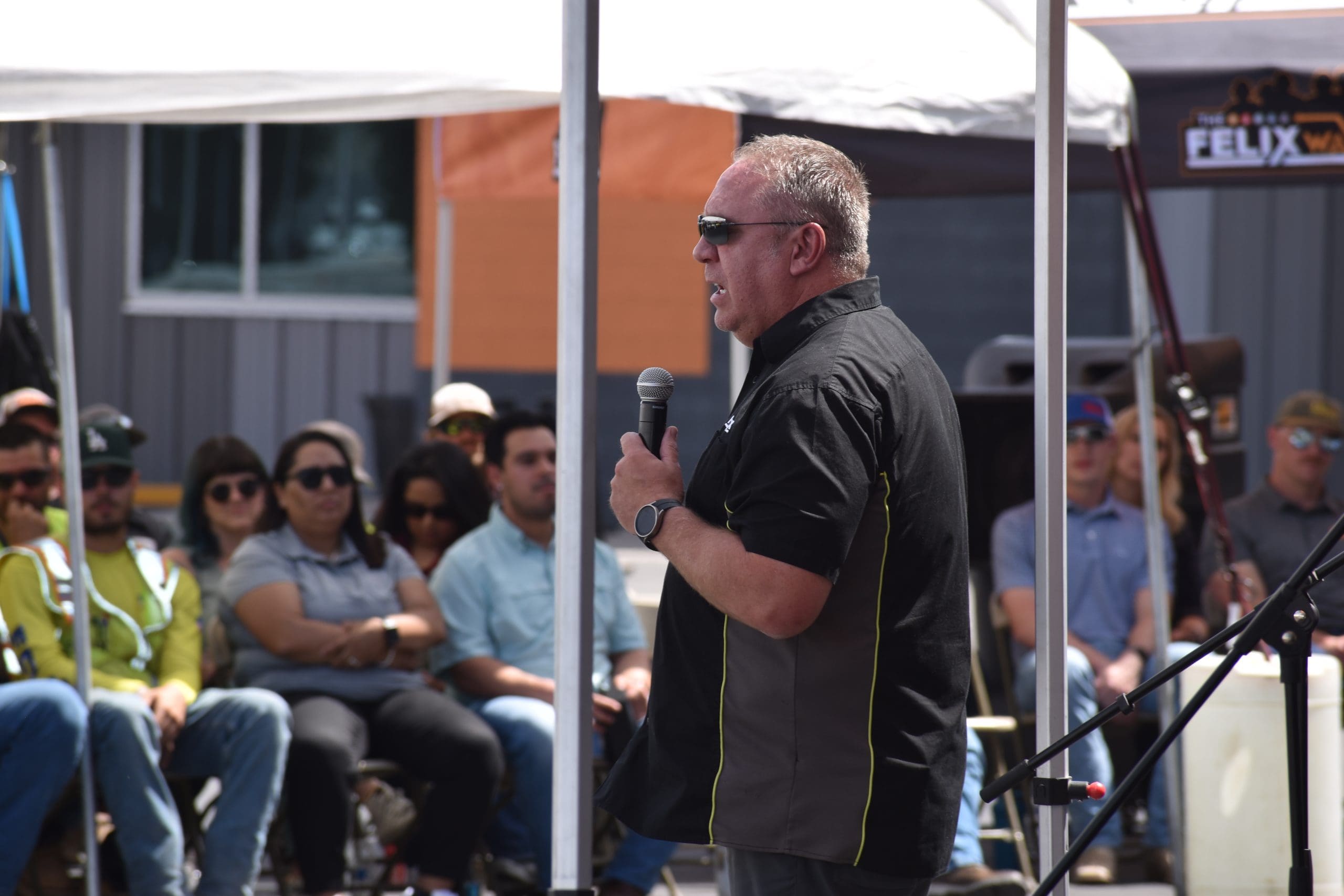A man speaks into a microphone while standing outdoors under a tent, addressing a seated audience.