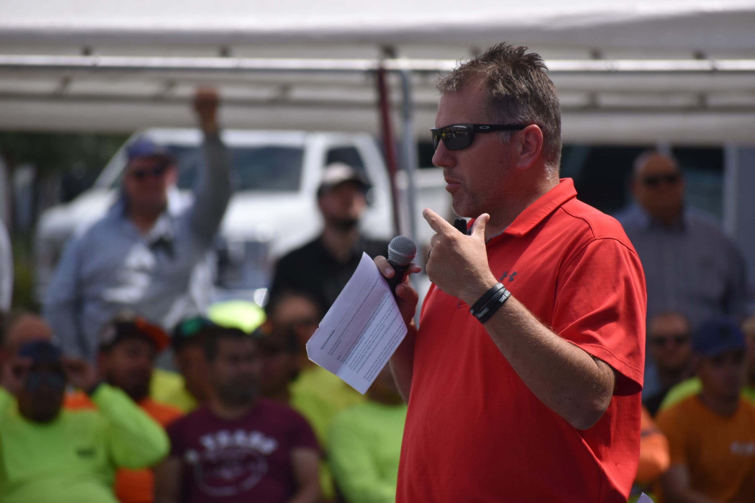 A man in a red shirt and sunglasses speaks into a microphone while holding papers; people sit under a canopy in the background.
