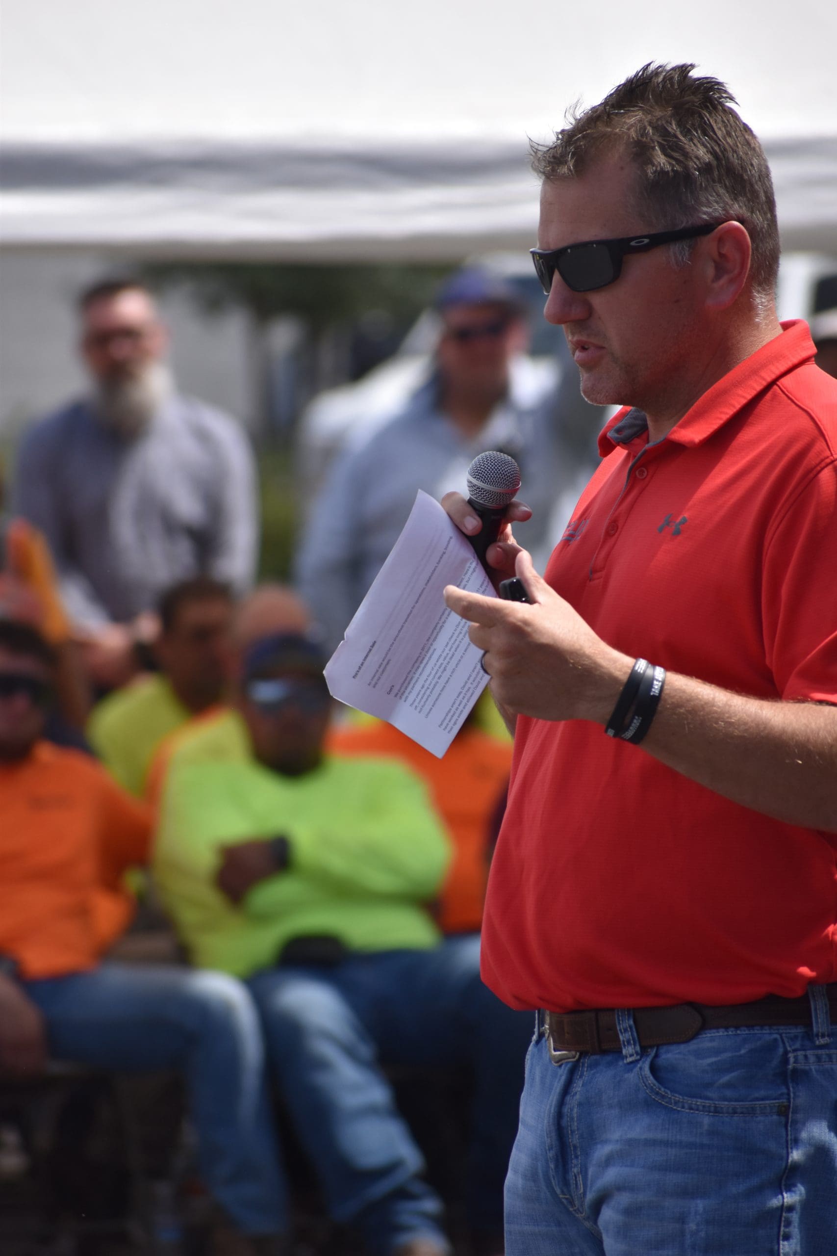 A man in a red shirt and sunglasses speaks into a microphone while holding papers; a group of people in safety clothing sit in the background.