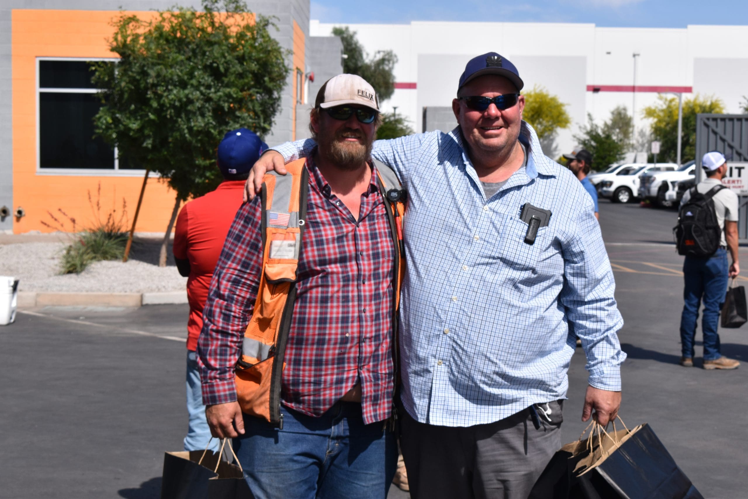 Two men stand outdoors in a parking lot, smiling with arms around each other, holding black gift bags; people and parked vehicles are visible in the background.