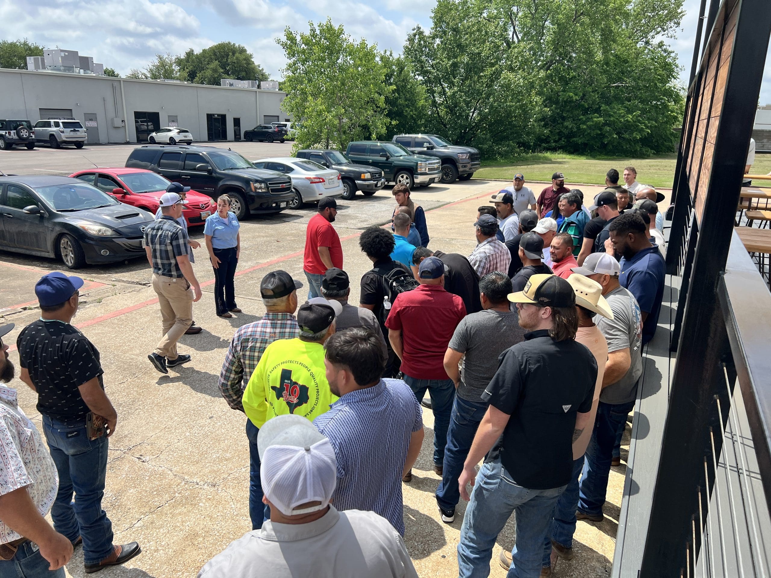 A group of people stands outside in a parking lot, listening to two individuals positioned at the front, near parked vehicles and a building.
