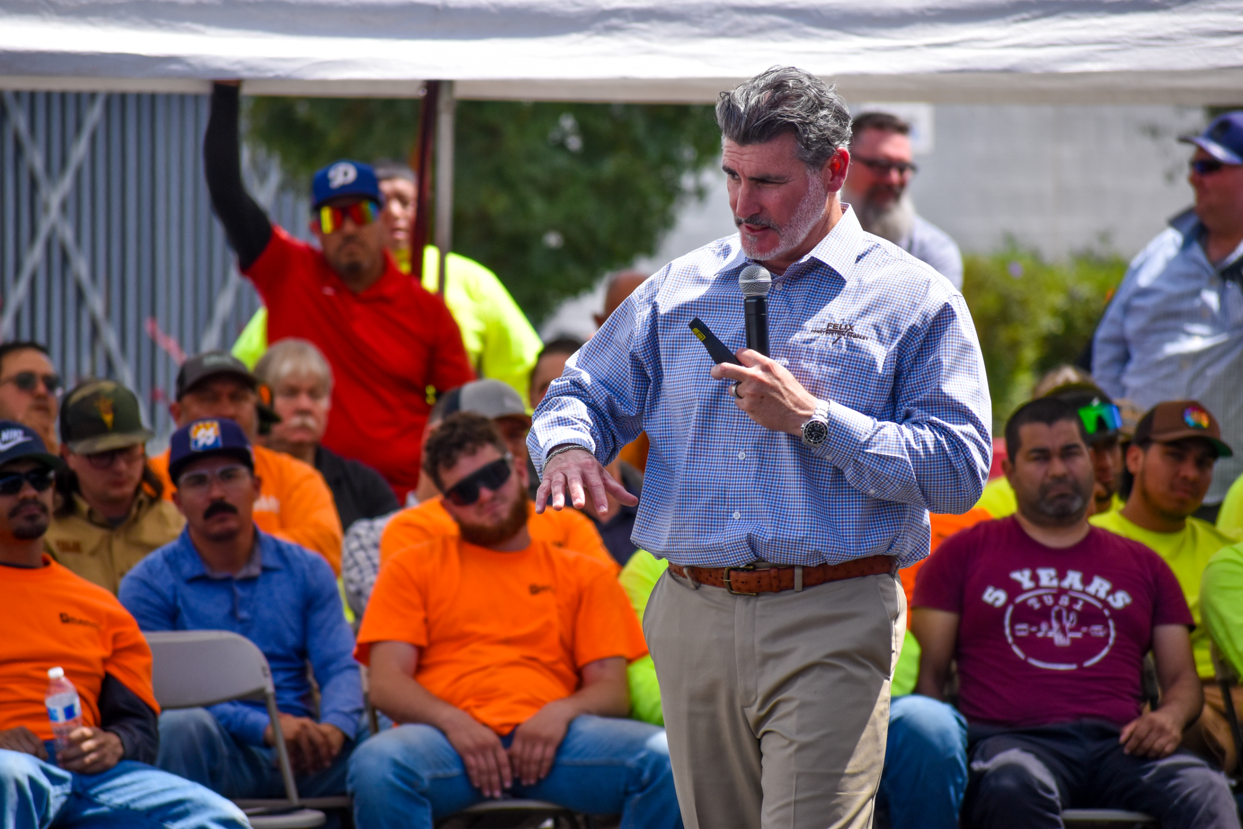 A man speaks into a microphone in front of a seated crowd of workers, some wearing bright safety shirts, at an outdoor event.