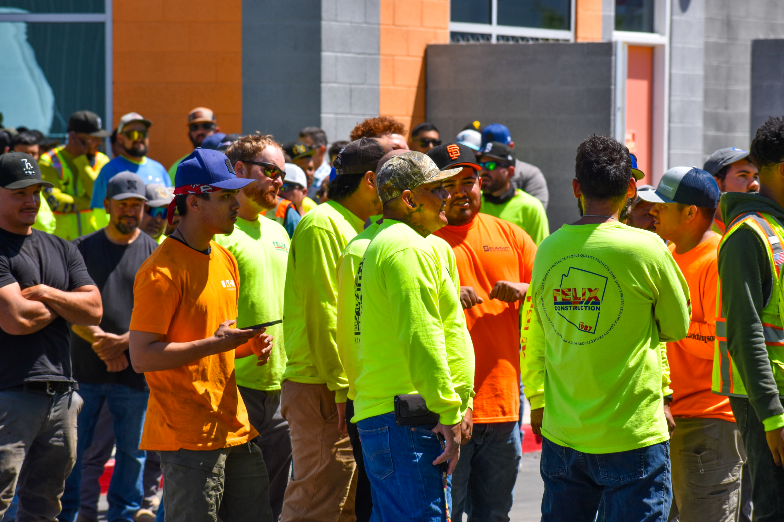 A group of construction workers in bright safety shirts and helmets gather and talk outside a building under construction.