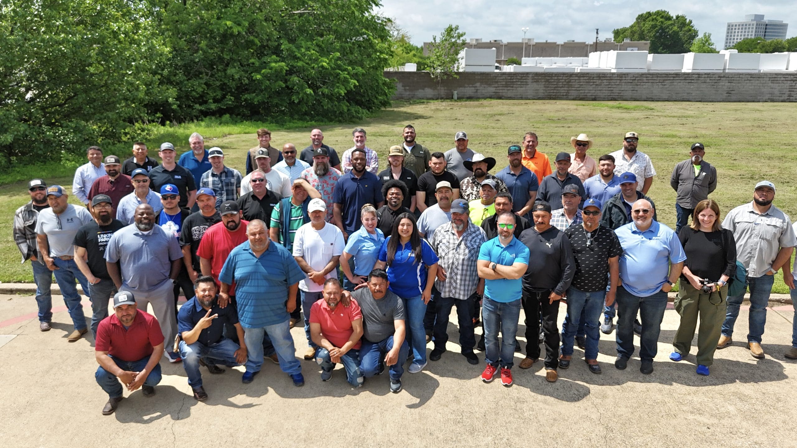 A large group of adults, mostly men, pose together outdoors on a paved area with grass and trees in the background.
