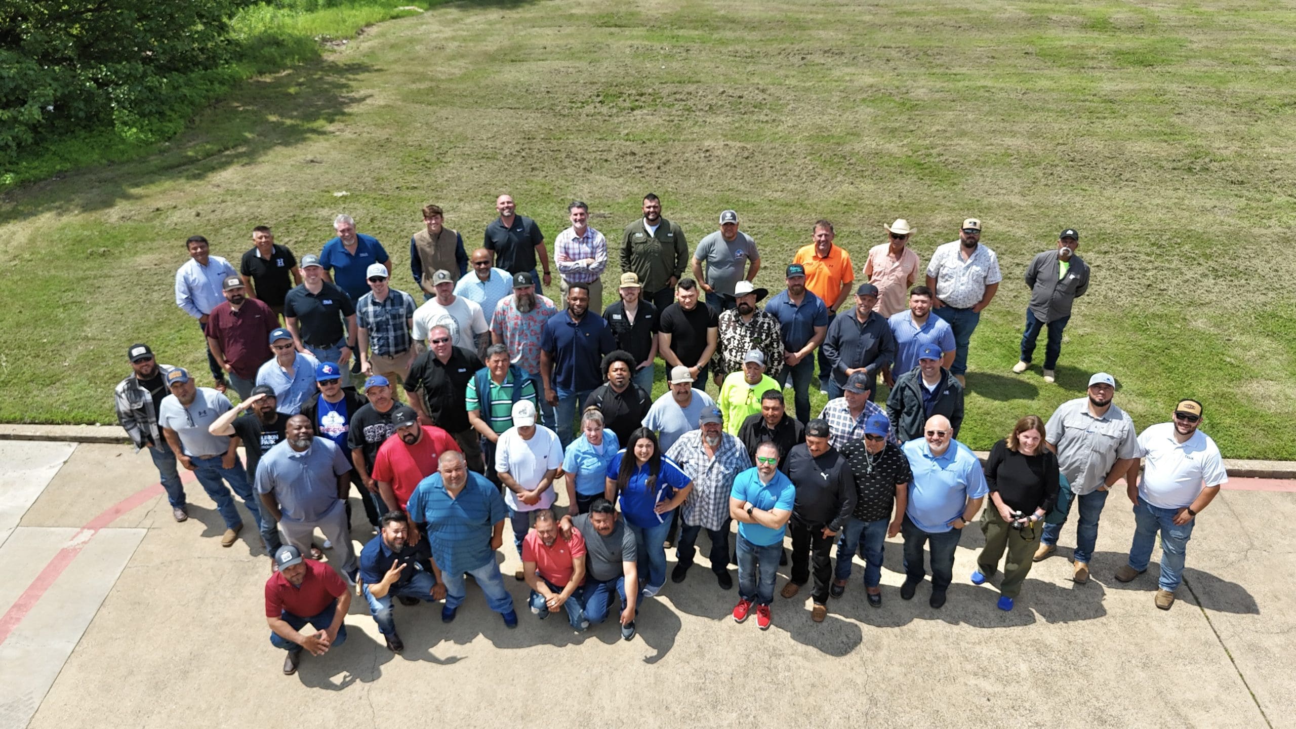 A large group of people, mostly men, pose outdoors on a concrete area next to a grassy field, facing the camera.